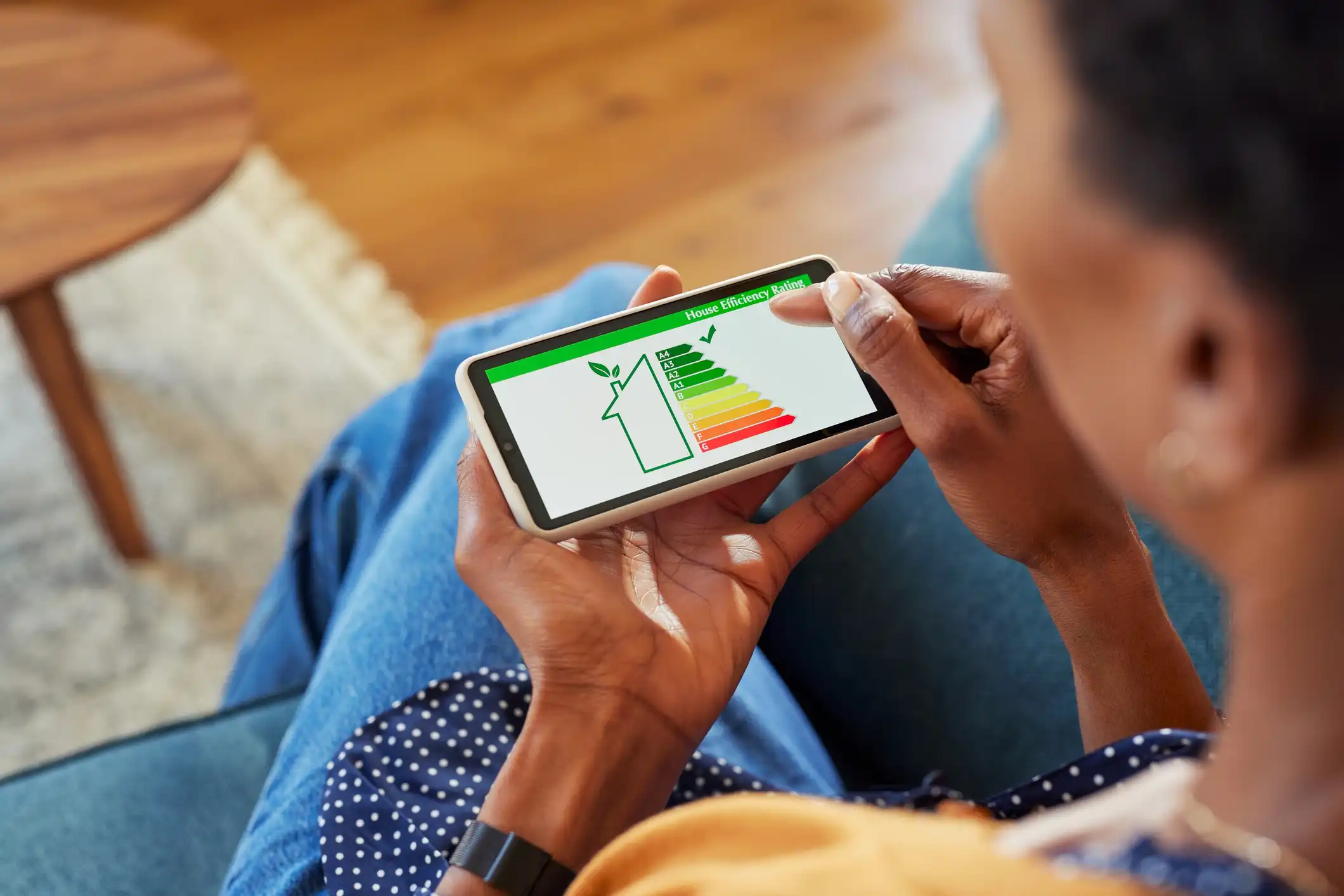A person with dark skin is sitting on a couch, holding a smartphone horizontally. The phone's screen displays a "House Efficiency Rating" graphic. The graphic features an outline of a house with a leaf inside, next to a vertical energy efficiency scale from A (dark green, most efficient) to G (red, least efficient). A small finger is touching the screen near the green A and B ratings, suggesting interaction with the results. The person is wearing a mustard-colored top, blue jeans, and a black watch or fitness tracker. The background is a soft-focus living room setting with a wooden side table and a portion of a rug visible.