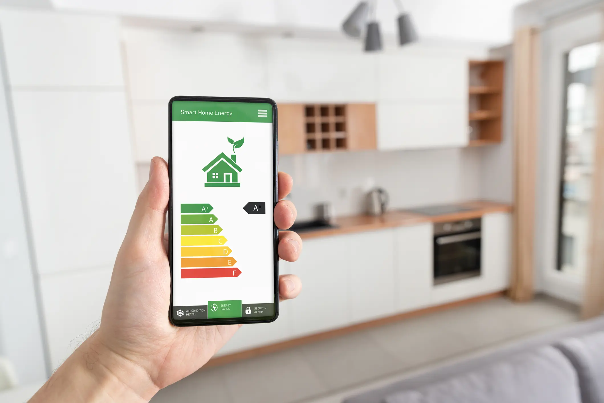 A person is holding a smartphone vertically in their right hand, foregrounded against a blurred, modern kitchen background. The phone displays a "Smart Home Energy" app.