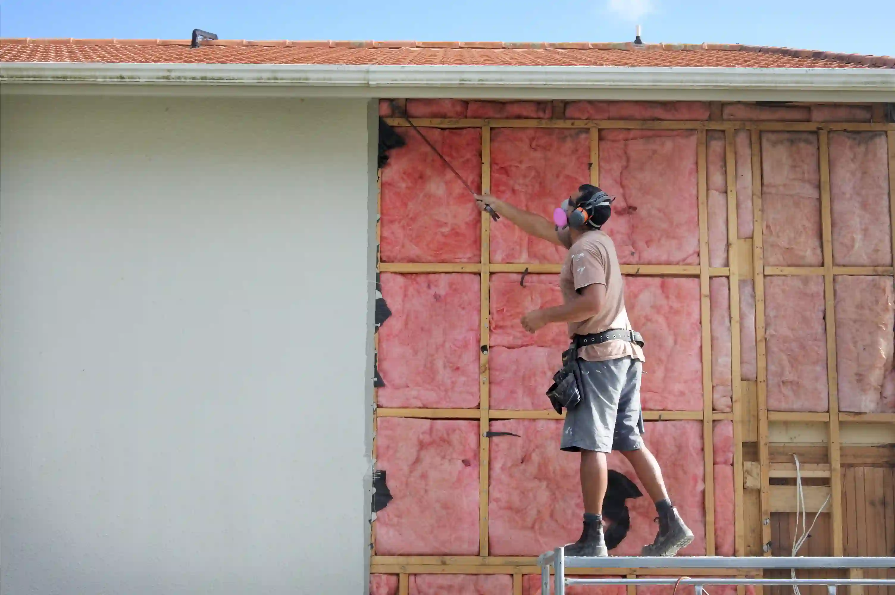 A male construction worker is standing on scaffolding next to the exterior wall of a house. The left side of the wall is covered in white stucco or siding, while the right side has been stripped, exposing the wooden stud framing and pink fiberglass batt insulation within the wall cavities. The worker is wearing a t-shirt, shorts, work boots, a tool belt, and personal protective equipment, including a respirator mask (or dust mask) and ear defenders.