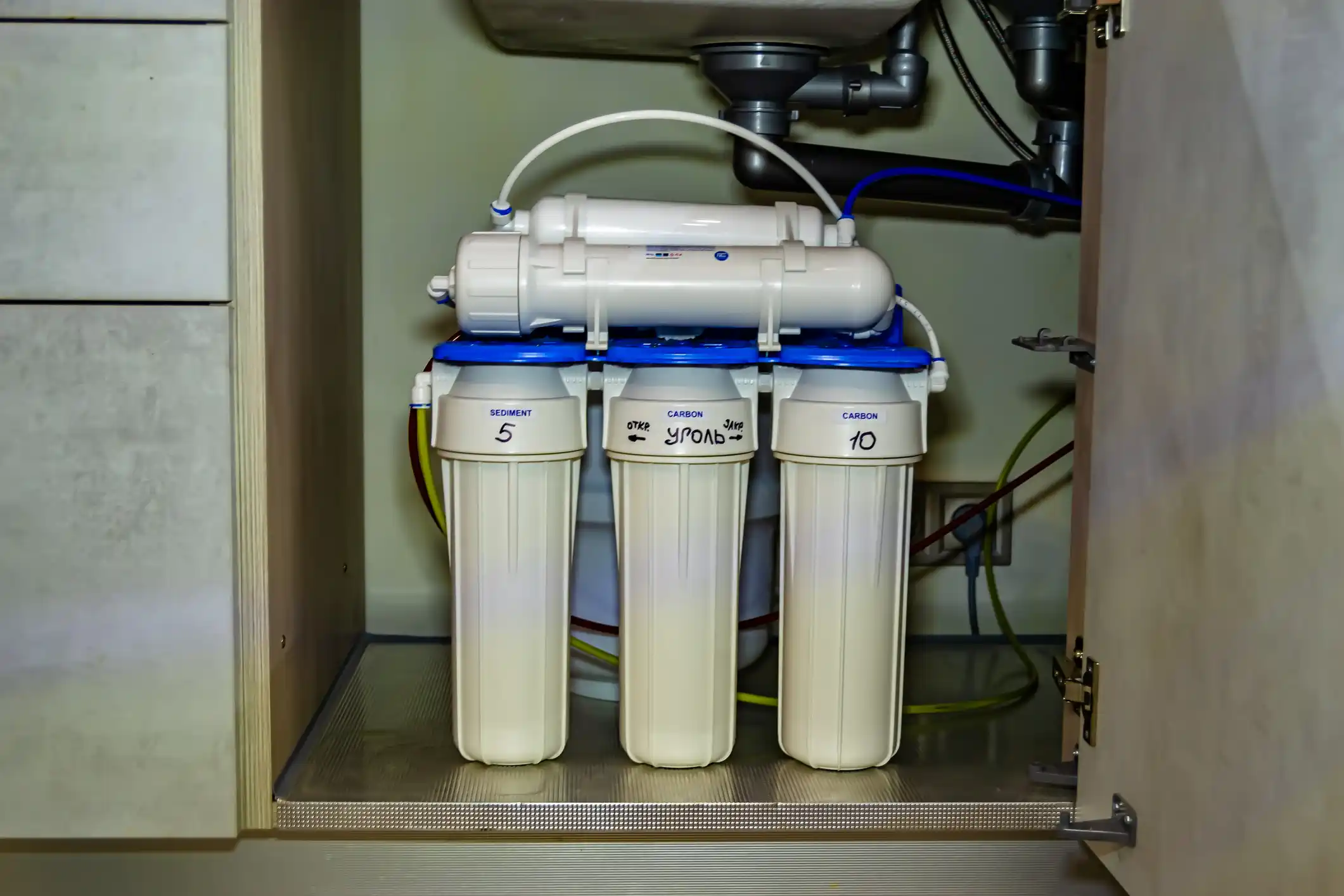 An under-sink reverse osmosis (RO) water filter system is installed beneath a kitchen sink. It features three vertical pre-filter canisters labeled Sediment, Carbon, and a third with Cyrillic writing