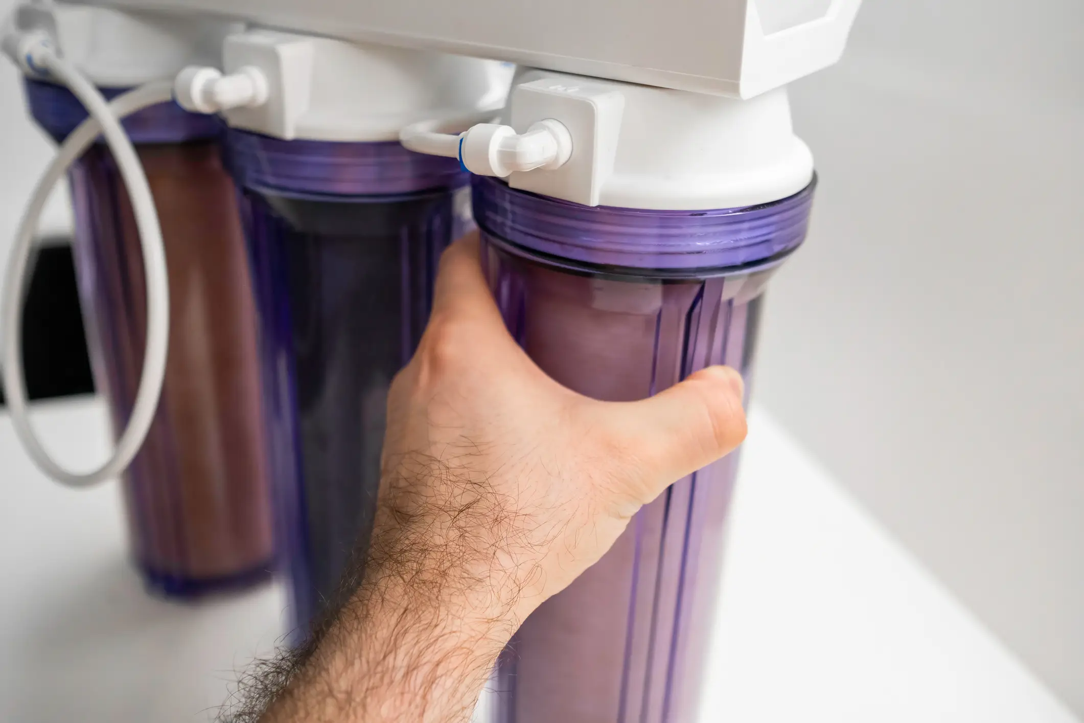 A person's hand is shown tightening or replacing a filter cartridge in a three-stage water filtration system, likely the pre-filters for a reverse osmosis unit. The canisters have purple clear/translucent housing, allowing the brown-to-purple filters inside to be seen, indicating they are in use or need replacement.