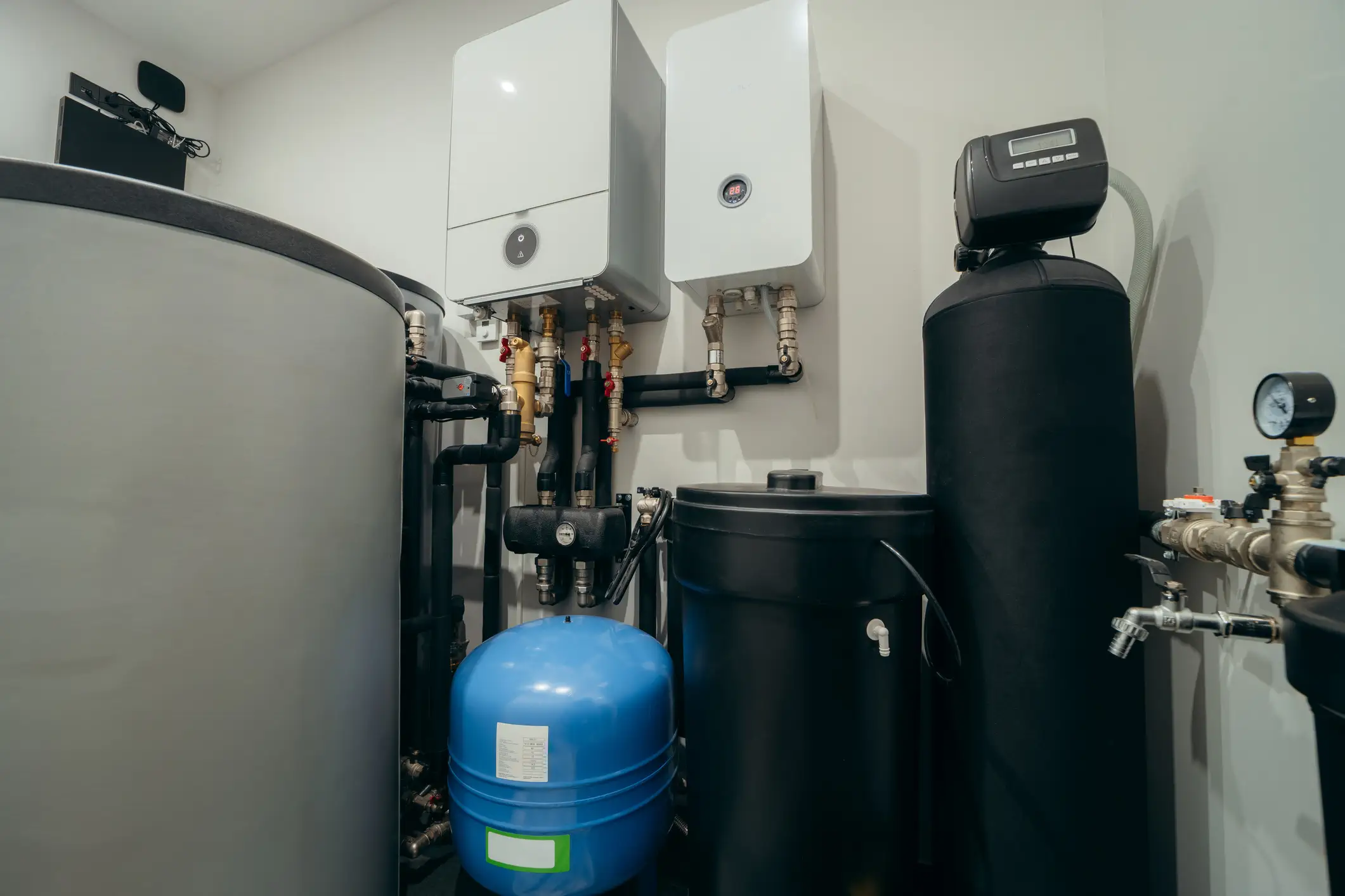 A complex home heating and water treatment system is installed in a utility room. It features two white wall-mounted boiler units, a large grey storage tank on the left, and a tall black cylindrical water softener tank