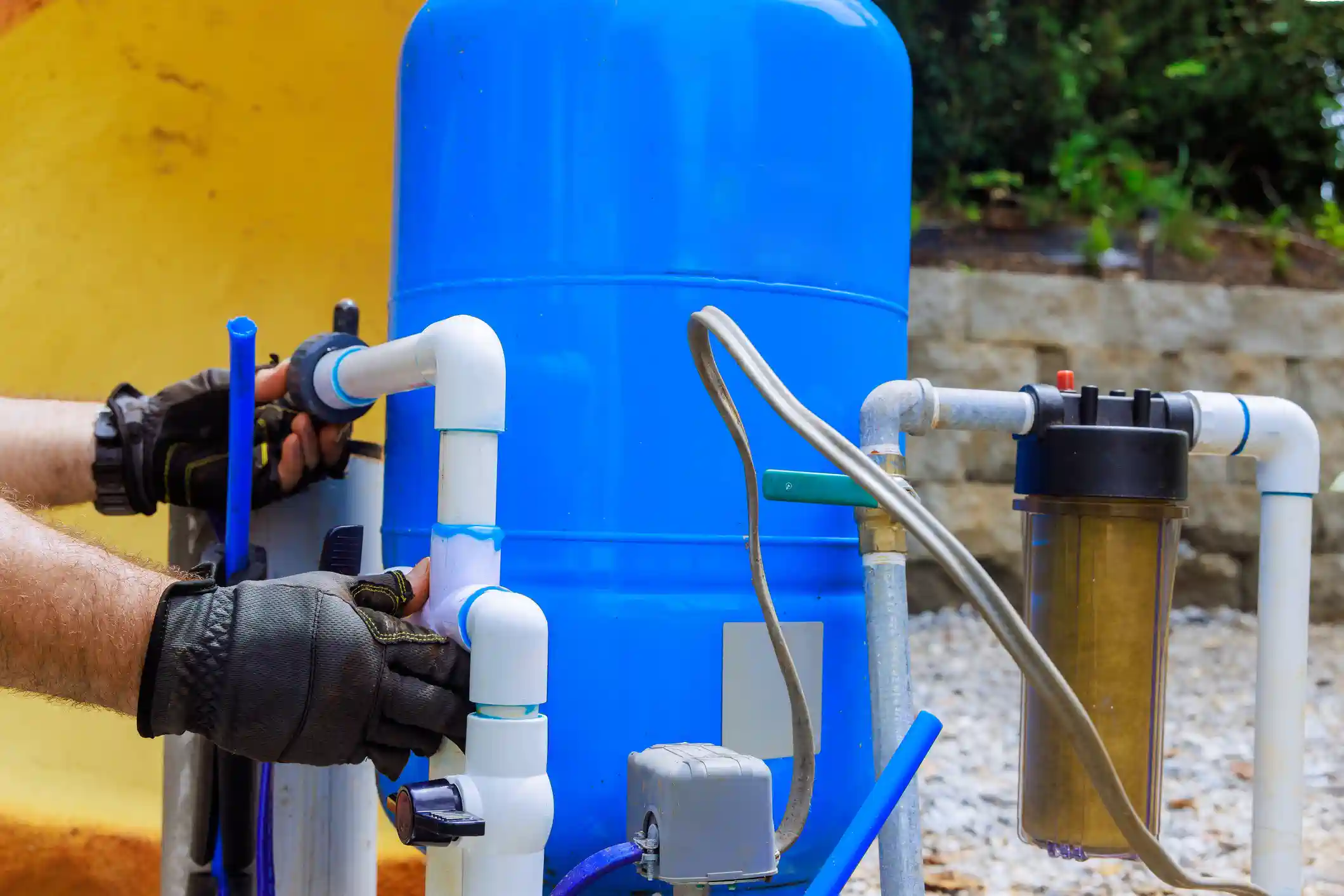 A pair of gloved hands is connecting white PVC pipes to a large, bright blue pressure tank, likely for a well water system, outdoors. A brown-tinted water filter canister and various valves and electrical connections are visible on the right, all set against a yellow wall and a gravel ground.