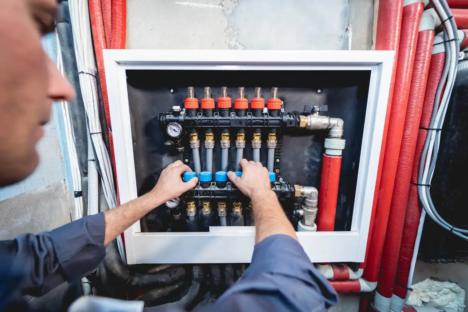 Plumber adjusting a heating manifold in a cabinet with red insulated pipes.