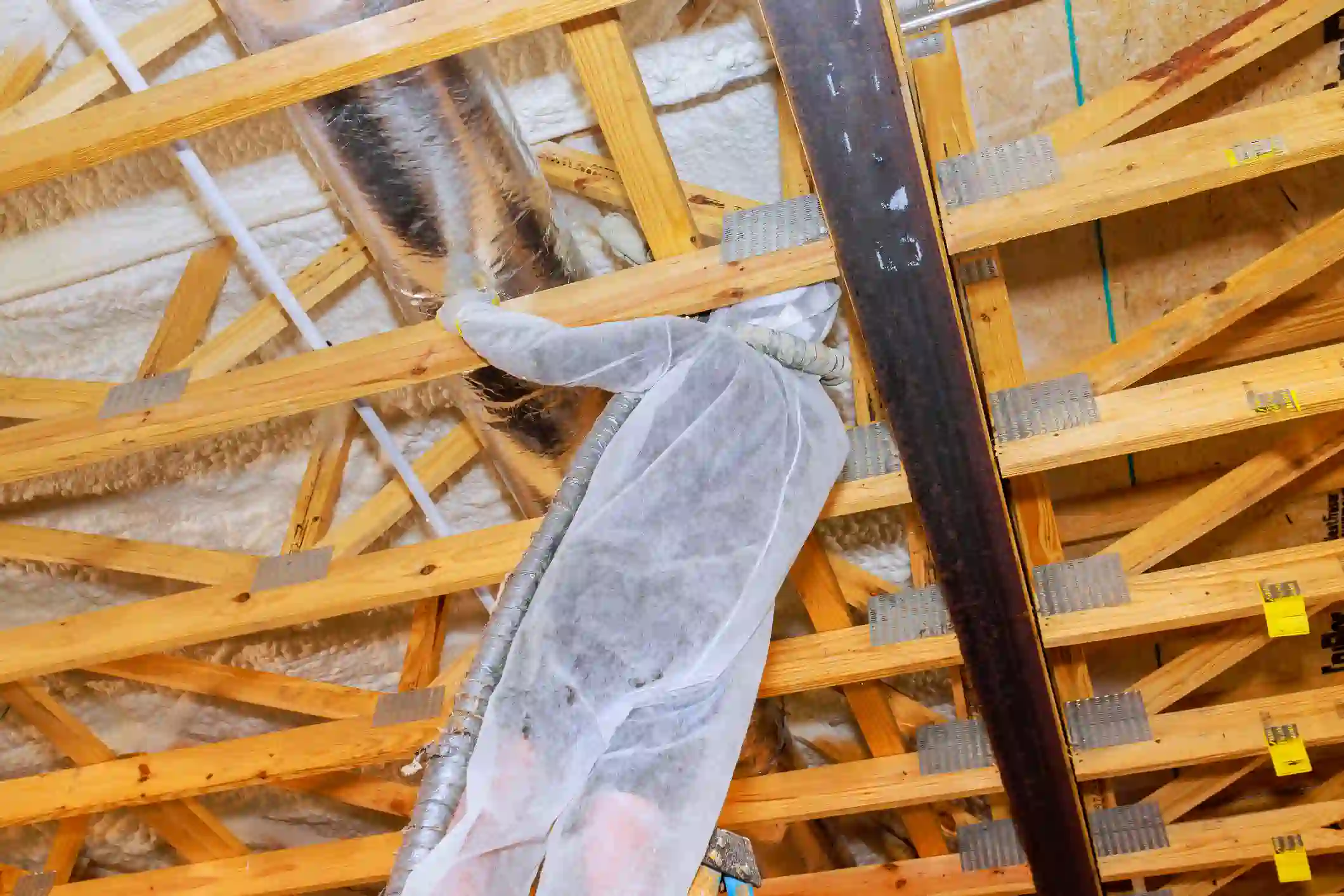 A construction worker wearing a white protective suit is installing spray foam insulation in an attic or vaulted ceiling. The worker is surrounded by exposed wooden trusses and is extending their arm with a hose and nozzle to apply the foam around a large, silver metal duct in the rafters.