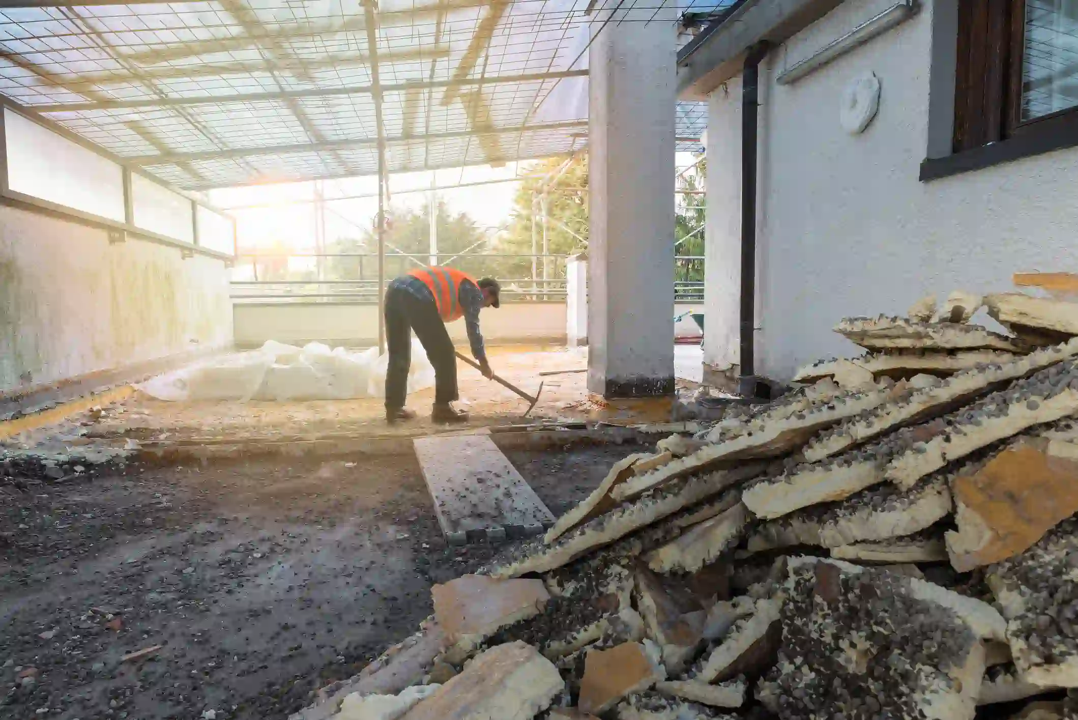 An outdoor scene showing construction or demolition work on a flat roof or covered patio area. In the foreground, there is a large pile of old, rigid insulation boards or roofing material that has been removed, consisting of layered light and dark materials. A worker, wearing a dark shirt, pants, a dark baseball cap, and a highly visible orange safety vest, is bent over in the mid-ground.