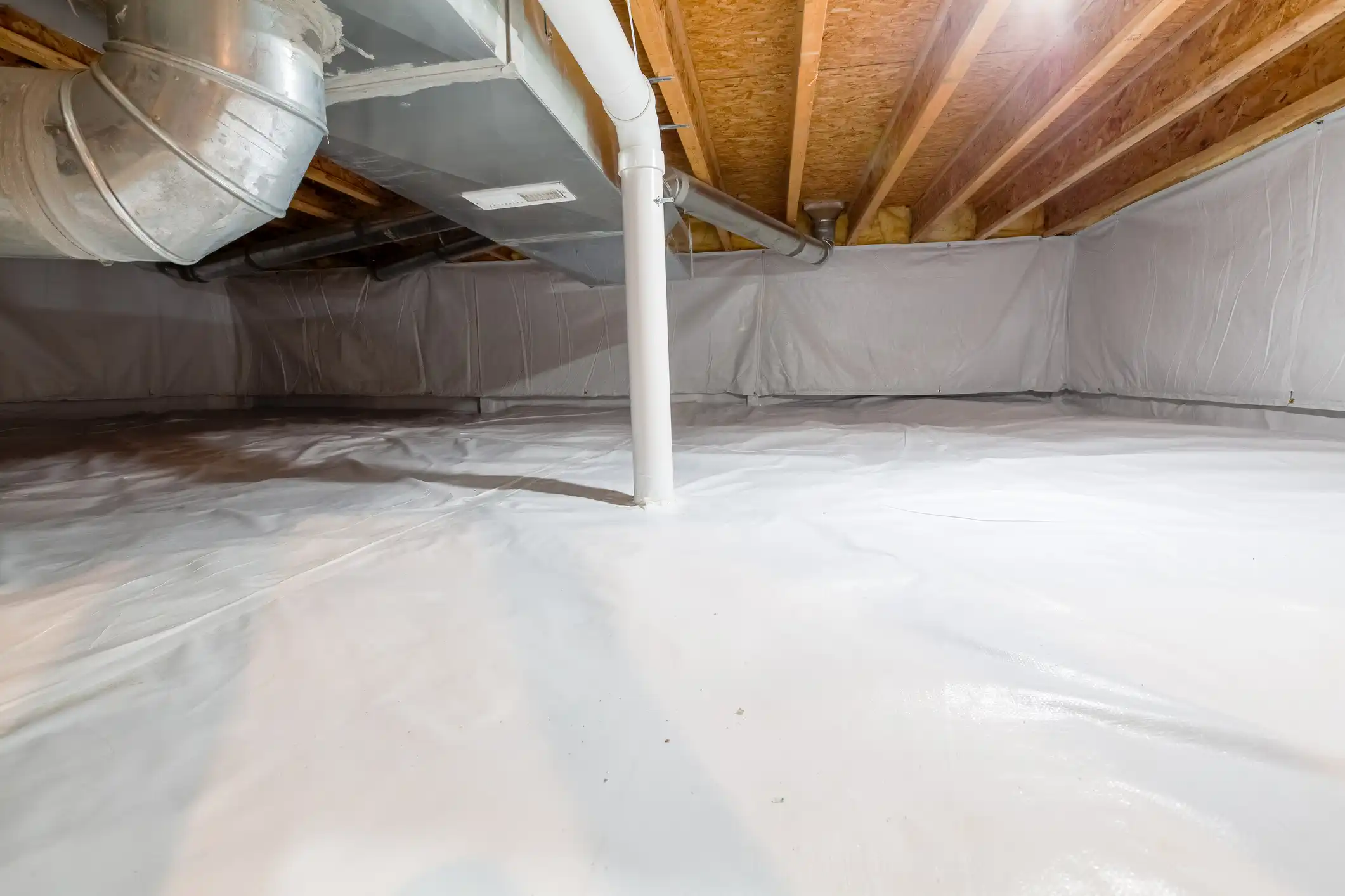  An image showing a well-lit, encapsulated crawl space that has been modernized for moisture control. The floor and foundation walls are covered with a continuous, clean white plastic vapor barrier (encapsulation liner). A white vertical support column stands near the center. Above, the underside of the floor joists and subfloor is visible, along with extensive HVAC and plumbing infrastructure, including a large, round galvanized metal duct, a square duct, and various white PVC pipes.