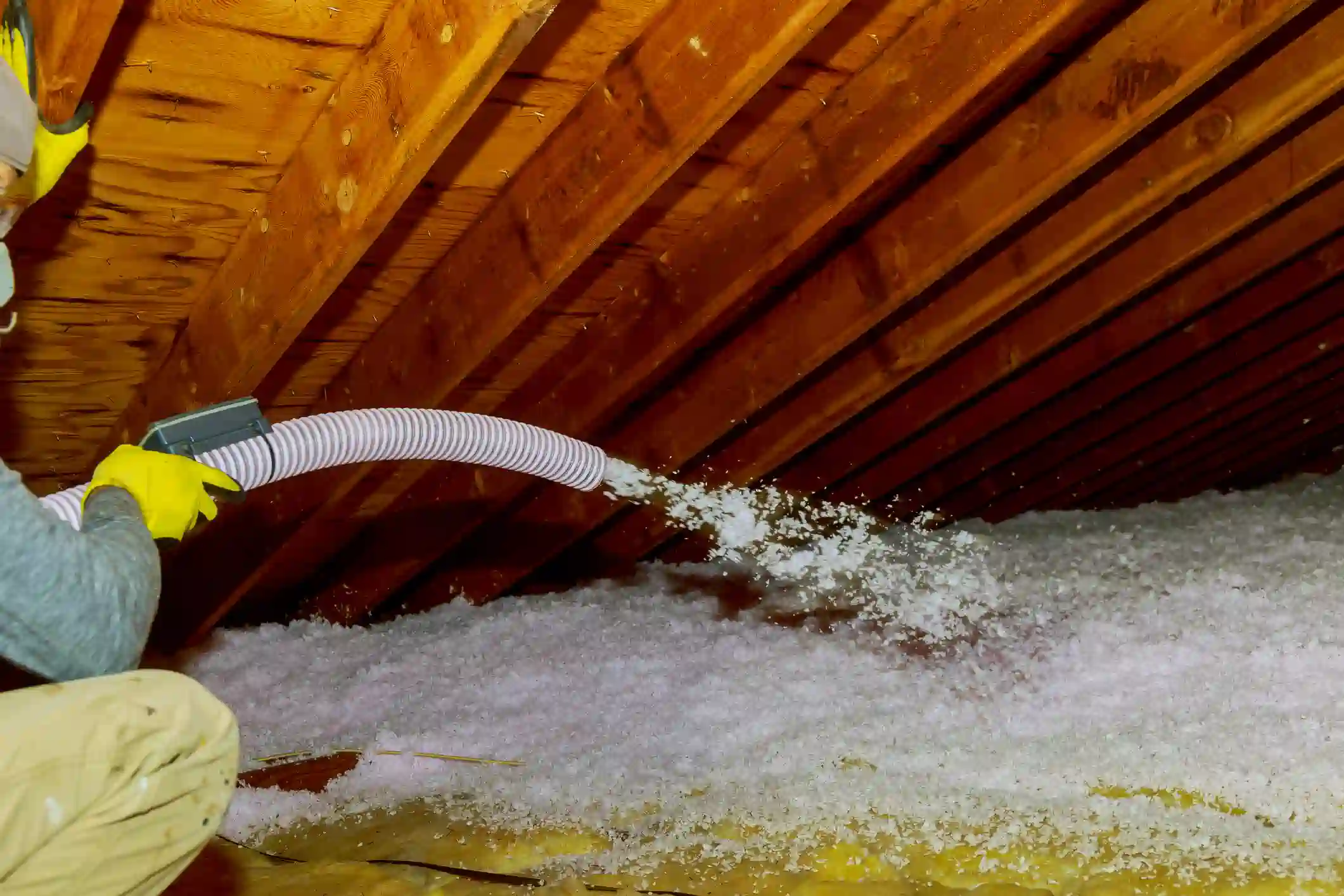 A low-angle shot in an attic shows a worker, partially visible on the left and wearing yellow gloves and goggles, operating a hose to install blown-in insulation. White, fluffy insulation material is being discharged from the end of the flexible, corrugated hose, covering the attic floor. The attic features exposed, warm-toned wooden rafters and ceiling boards above. A layer of yellowish material is visible beneath the newly applied white insulation.