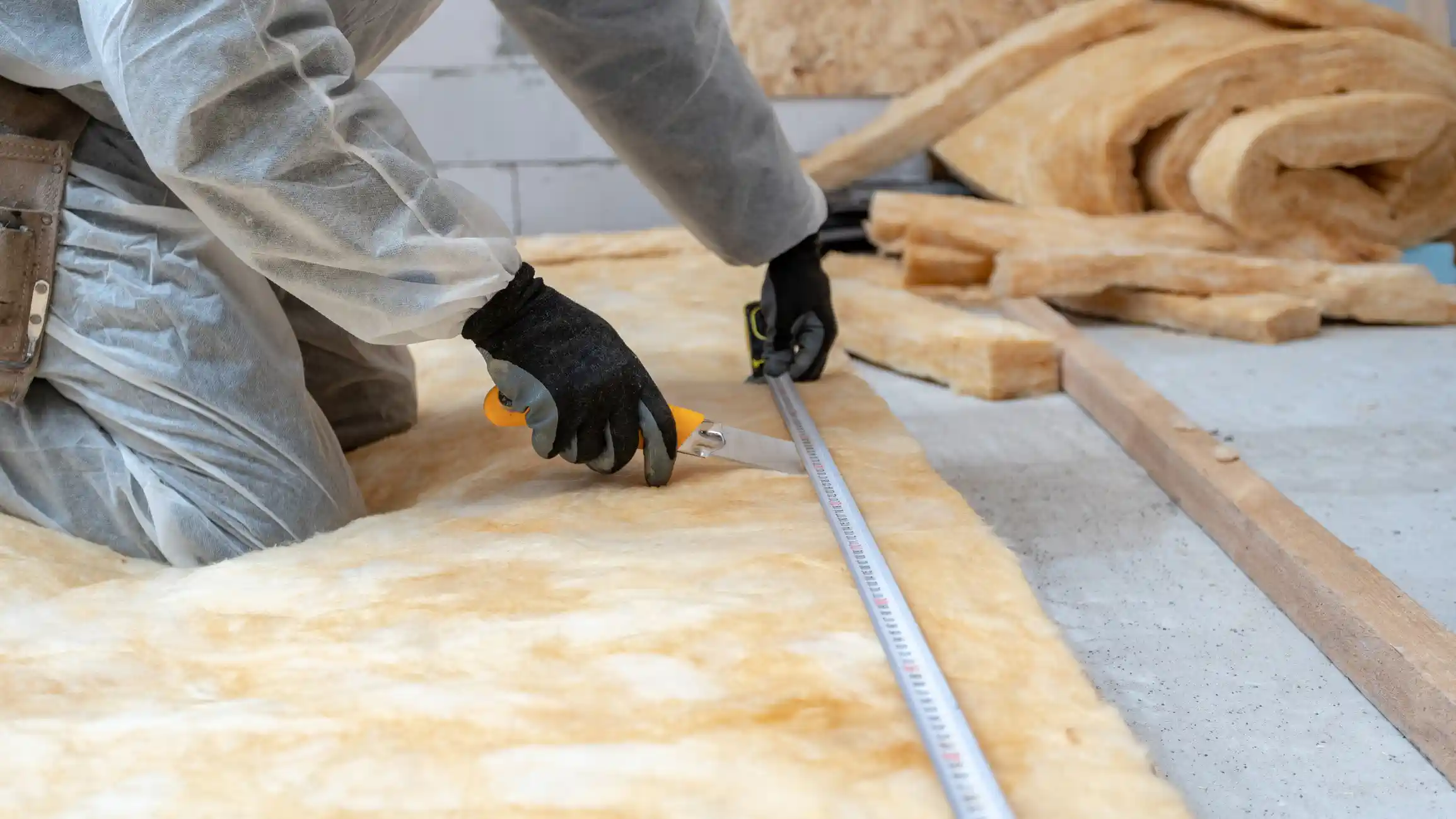  A construction worker, wearing a white protective suit and black gloves, is cutting a roll of fiberglass batt insulation on the floor. They are using a utility knife and a long metal tape measure to ensure a precise cut. Rolls of insulation and a wooden plank are visible in the background.