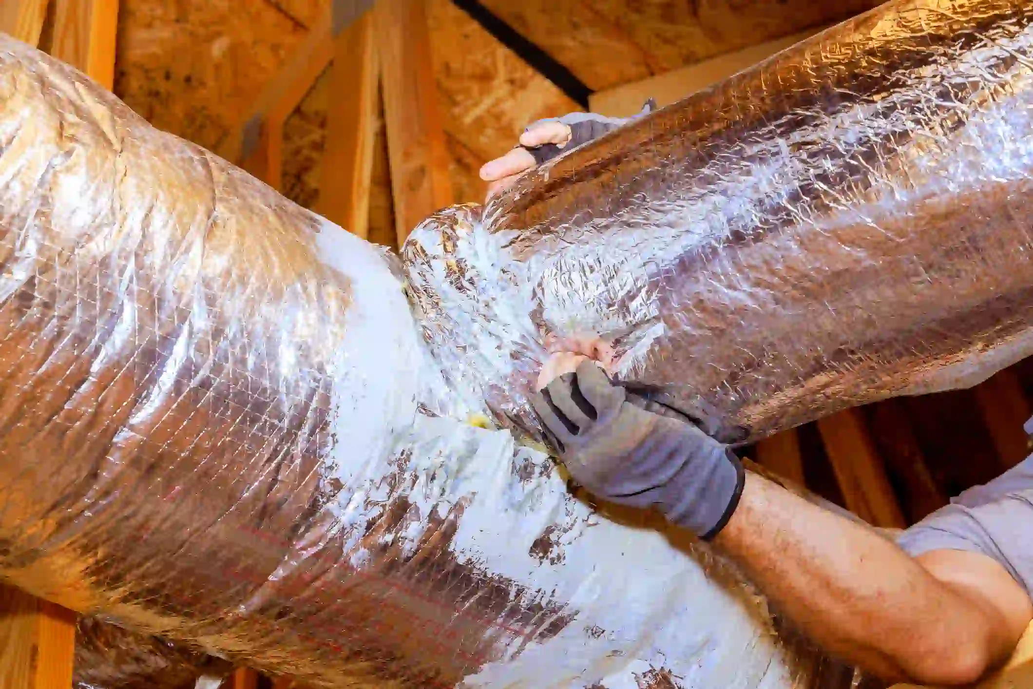  A worker's gloved hands are shown applying white mastic sealant to the joint of two pieces of flexible, foil-insulated HVAC ductwork in an attic or crawlspace. The background features exposed wooden roof rafters or joists.
