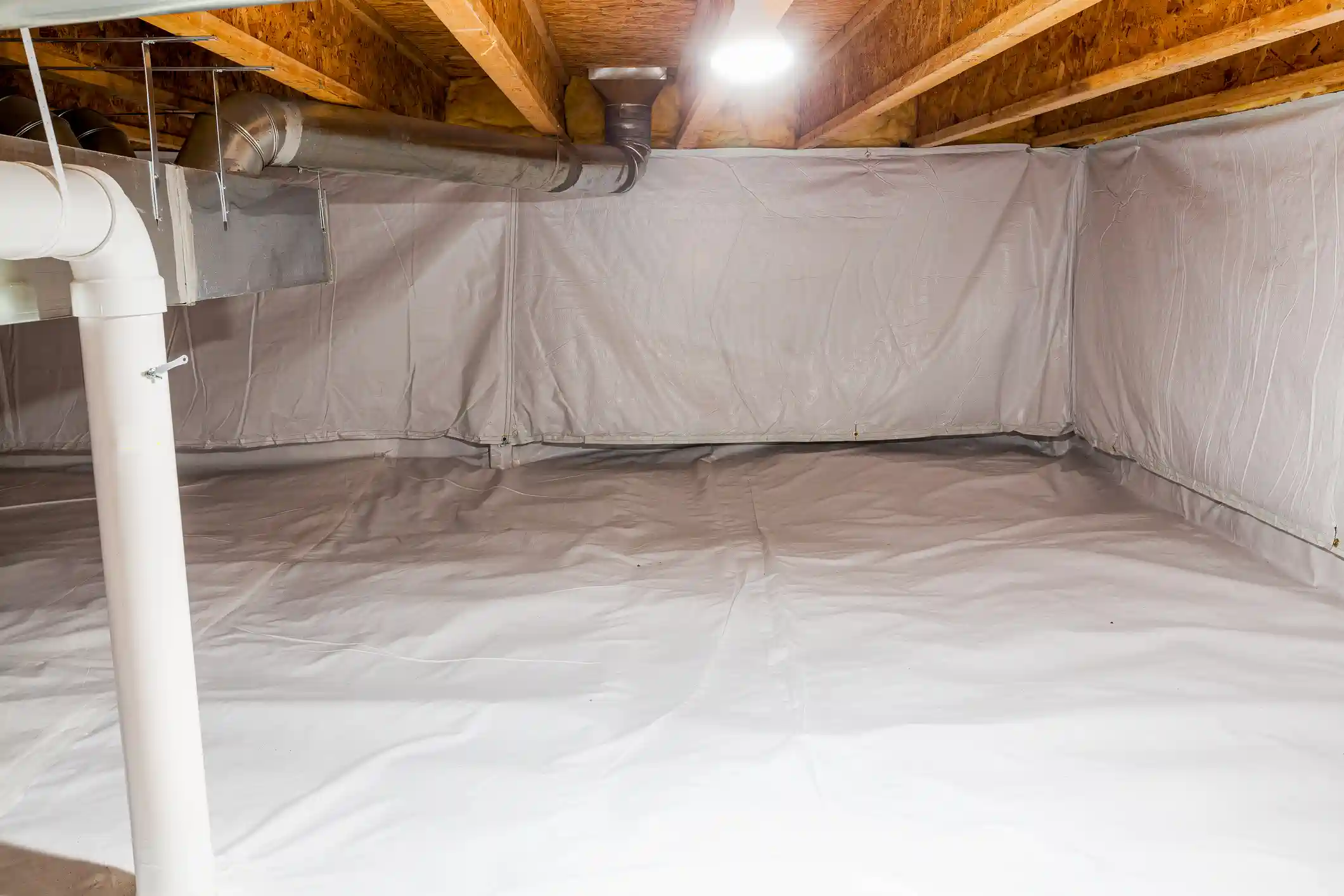 An image of a clean, encapsulated crawl space. The concrete floor and foundation walls are completely covered with a thick, continuous white plastic vapor barrier (encapsulation liner). Above, the wooden floor joists and OSB subfloor are visible, with some fiberglass insulation tucked into the rim joists. White PVC plumbing pipes, a large galvanized metal duct, and a square ventilation boot run along the ceiling. The space is bright and illuminated by an overhead light.