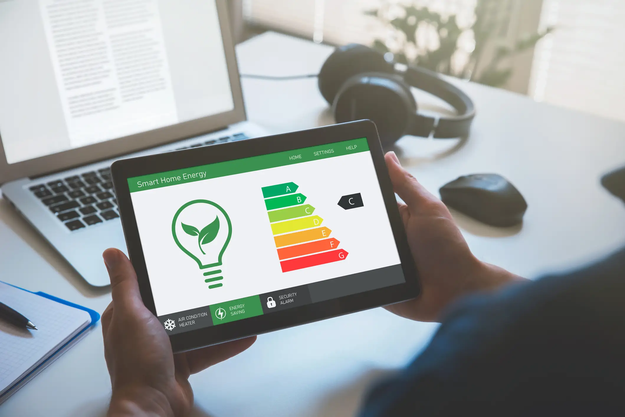 A person is holding a tablet horizontally, displaying an app called "Smart Home Energy." The screen shows a prominent graphic: a lightbulb with a leaf filament on the left, symbolizing eco-friendly energy, next to a colored energy efficiency scale from A (dark green, most efficient) down to G (red, least efficient). A black arrow points to the C level, indicating the home's current efficiency rating. Below the main graphic, there are icons for Air Condition, Energy Saving (currently highlighted), and Security Alarm. The background is a blurred office or home workspace with a laptop, headphones, a mouse, and a notebook.