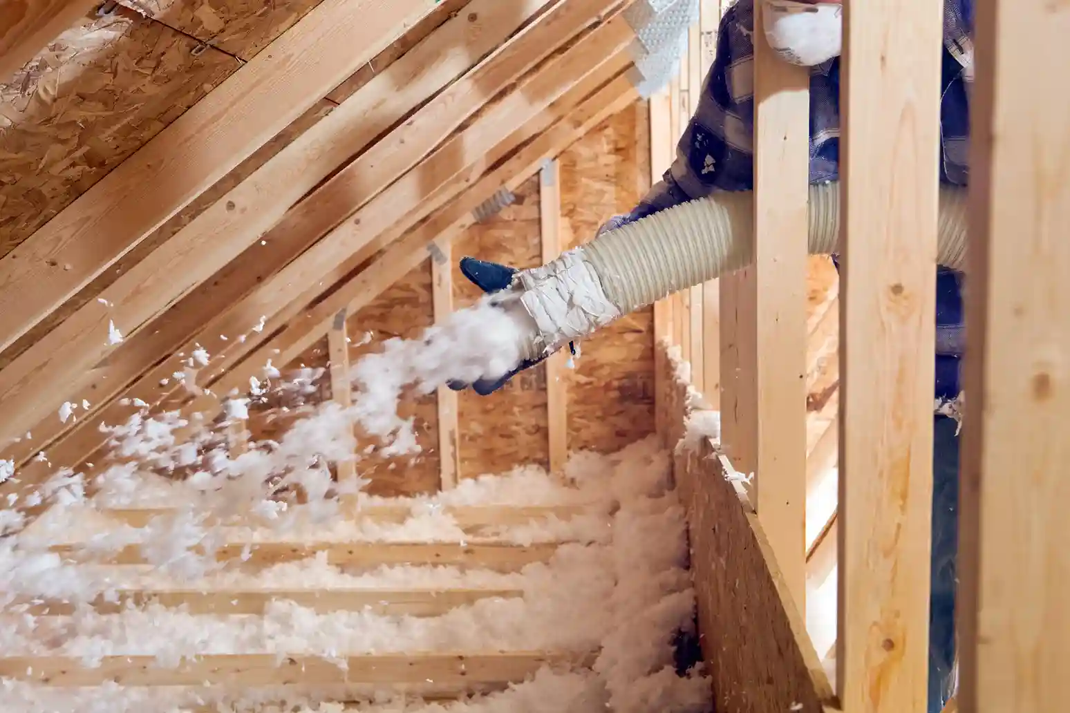 A person is using a large white hose to blow white, fluffy insulation material into the floor joists of an attic.