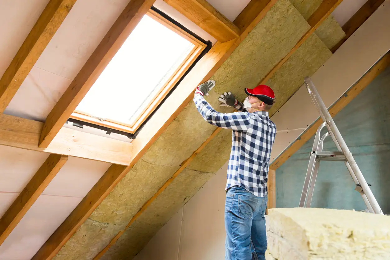 Technician in a plaid shirt installing thick mineral wool insulation batts between wooden rafters near a skylight