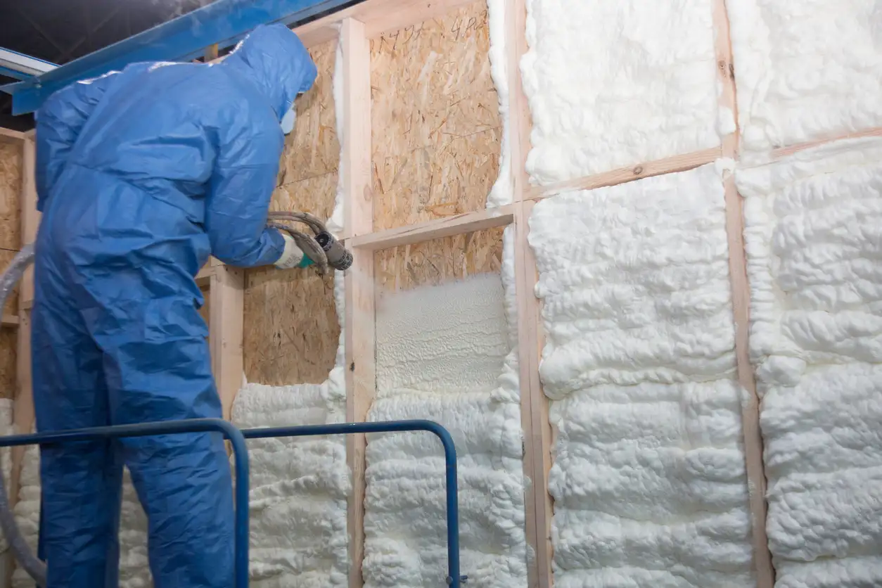 Professional in a blue protective suit spraying white expanding foam insulation into a residential wall cavity