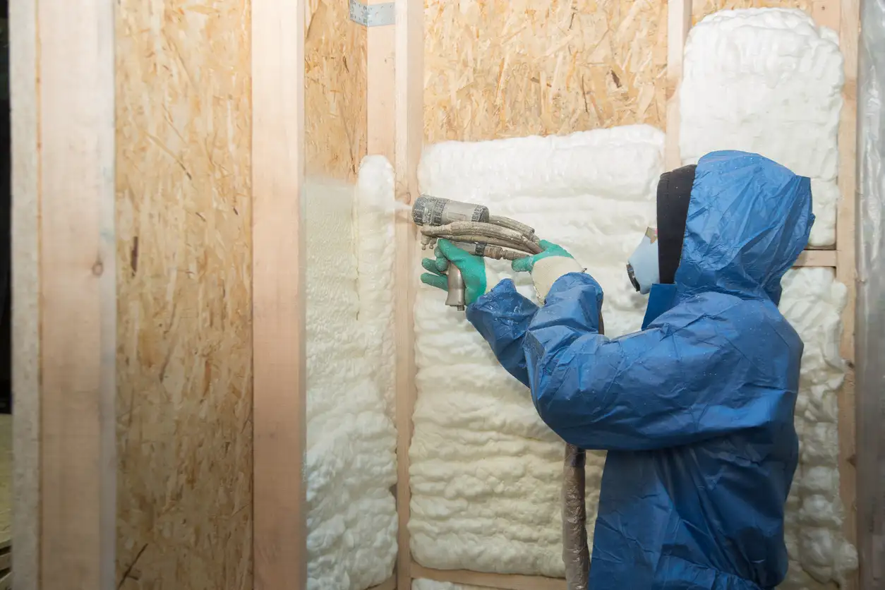 Installer in a blue hard hat placing yellow fiberglass insulation batts into a wooden wall frame
