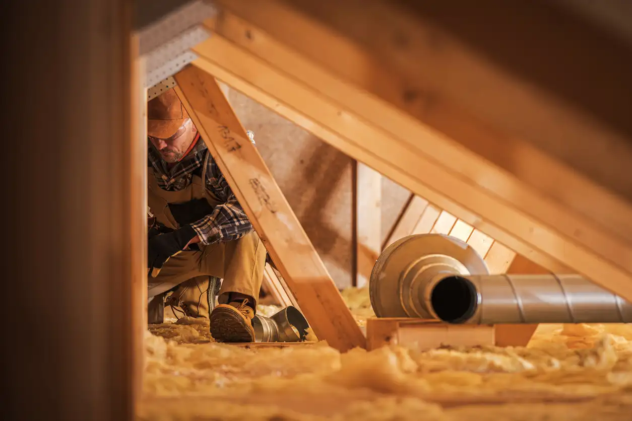 Professional installer working in a cramped attic space near metal ductwork and yellow fiberglass batt insulation