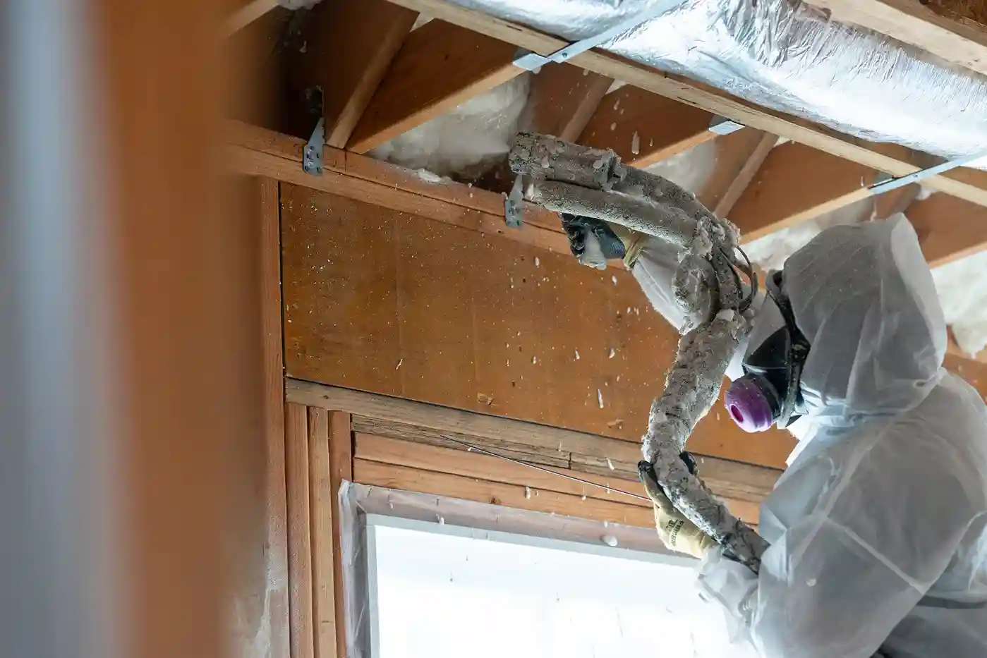 A technician in a full white suit and respirator is spraying foam insulation into the ceiling of an attic.