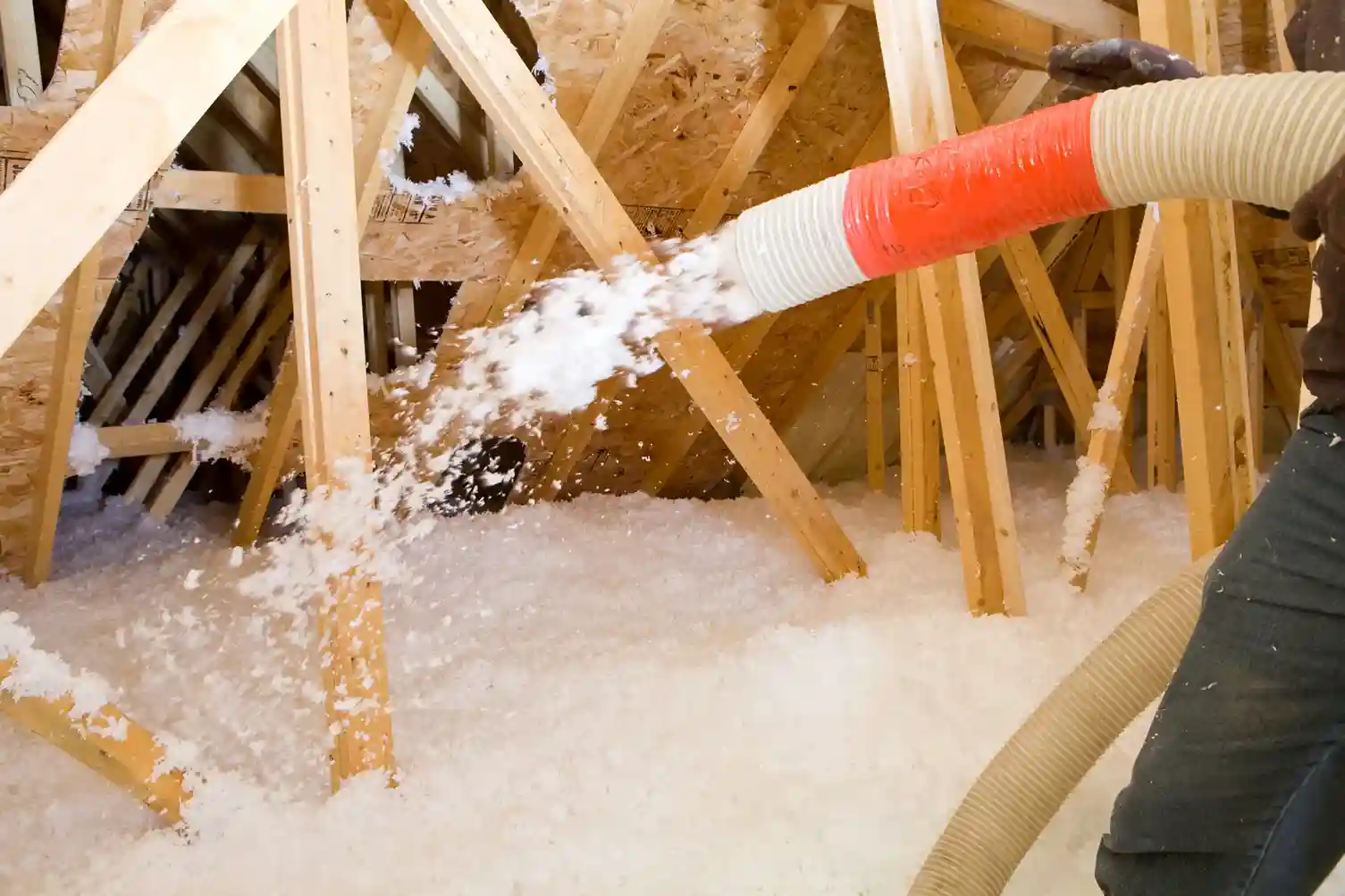 A person is using a large hose to spray white, fluffy insulation material onto the floor of an attic, with wooden beams in the background.