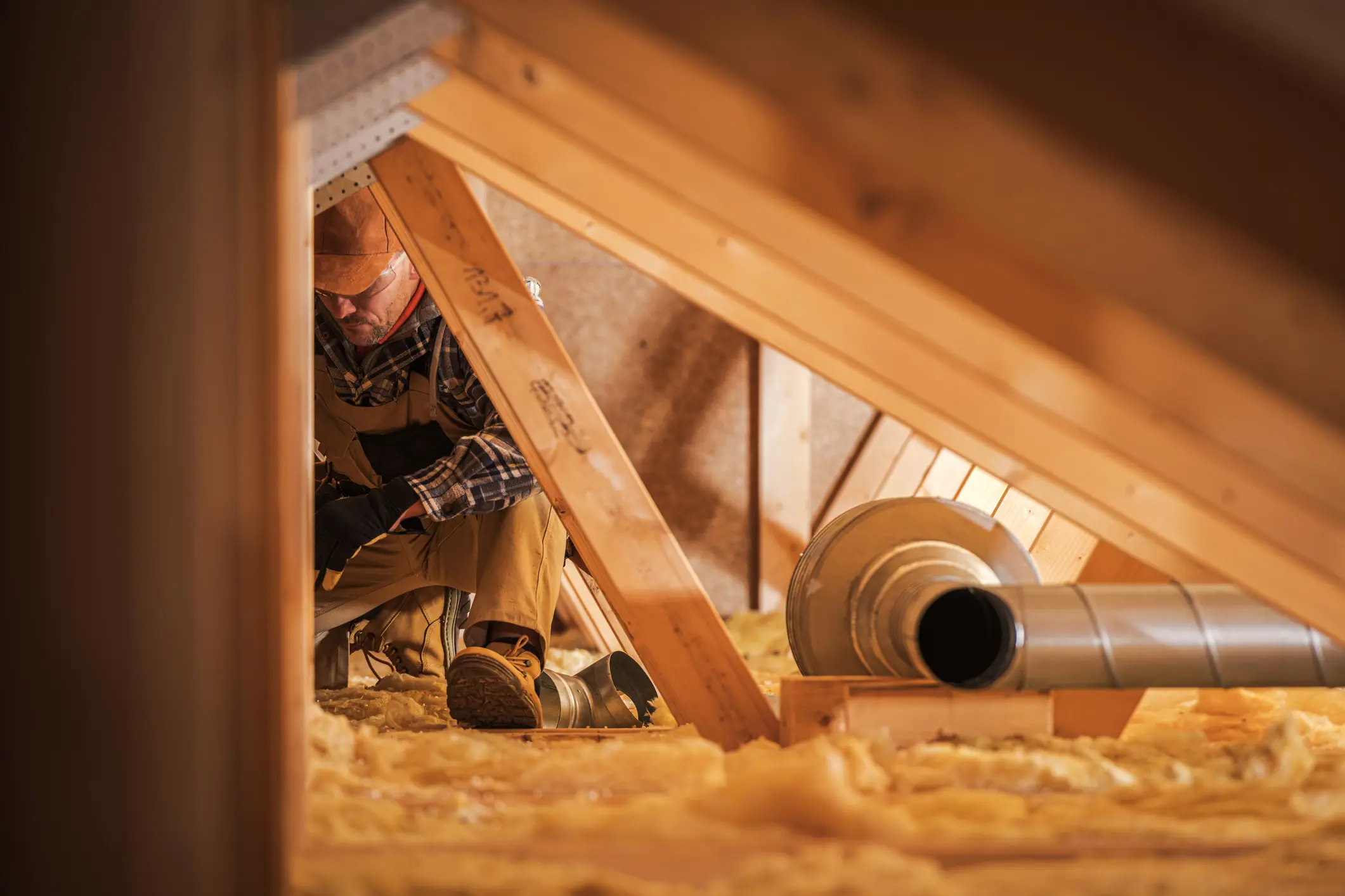 A low-angle shot shows a man in a cap and work clothes working on an HVAC or ventilation pipe in an attic with yellow insulation.