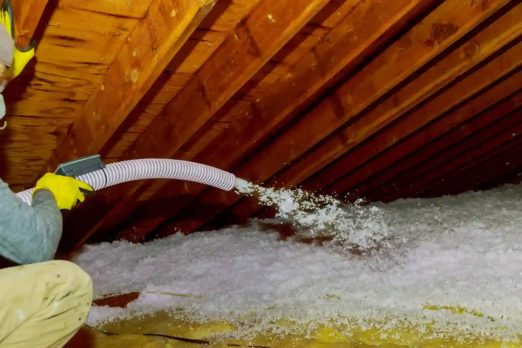 Worker in yellow gloves using a large hose to blow white loose-fill insulation into a wooden attic