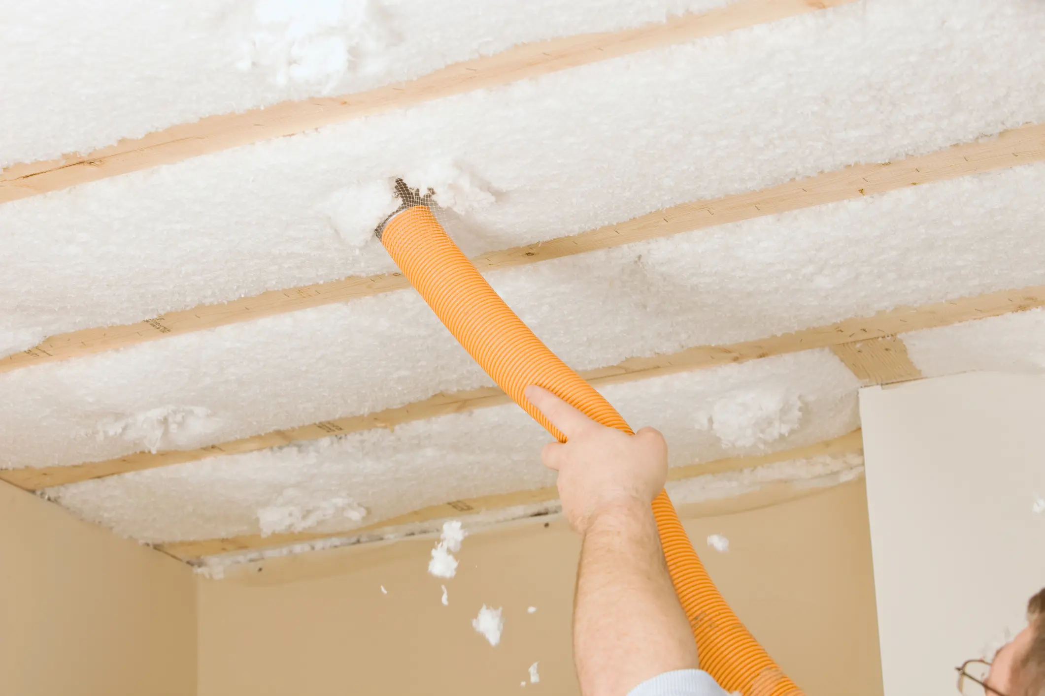A person uses an orange hose to install blown-in insulation into a ceiling joist space.