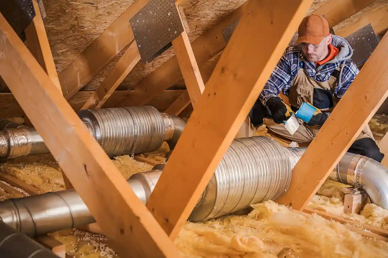 A man in work clothes and a cap is working on installing flexible silver ducting in an attic filled with yellow-colored insulation.