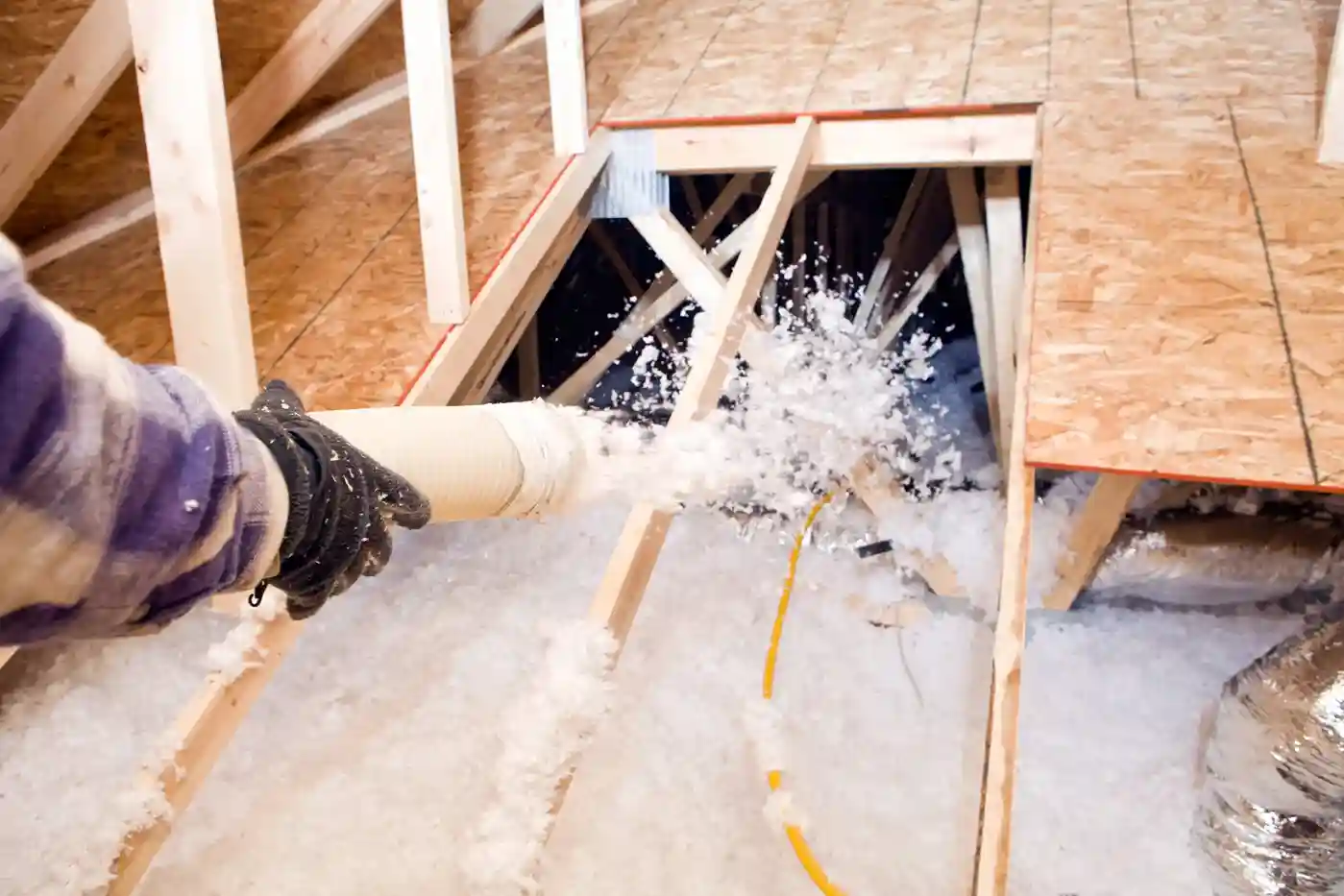 A person's hand holds a large hose, which is blowing white insulation into a narrow gap in a wooden attic frame.