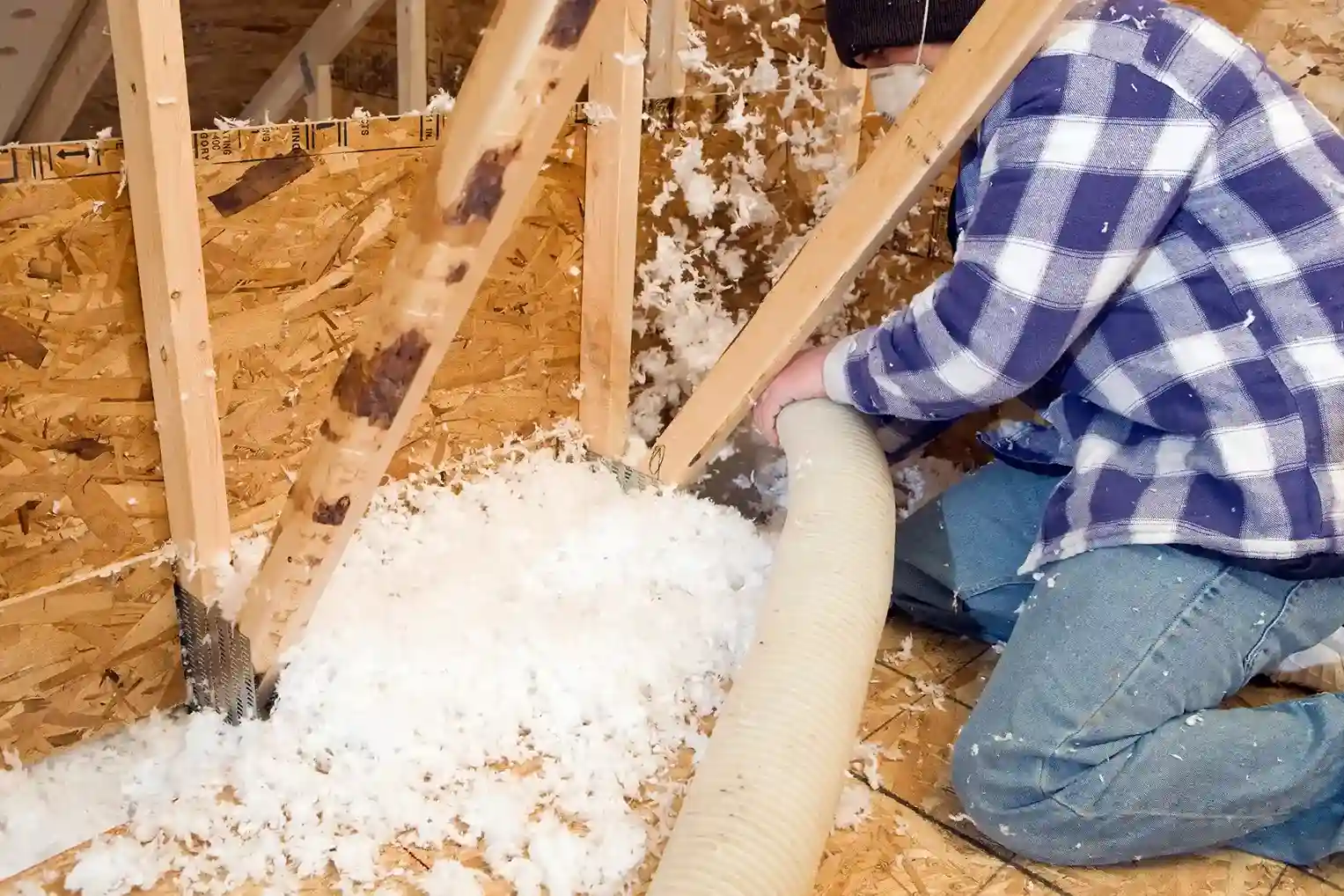 A person in a plaid jacket and jeans kneels in an attic, holding a hose to blow white insulation between wooden joists.