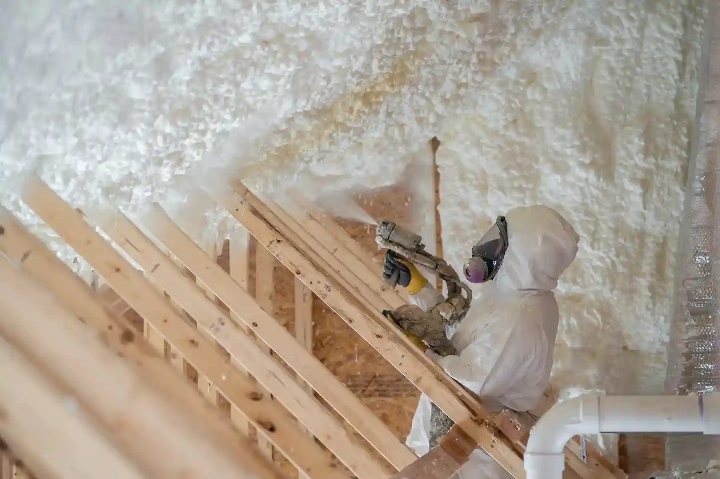 A person in a full protective suit and mask is using a spray gun to apply white foam insulation to the interior of an attic ceiling.