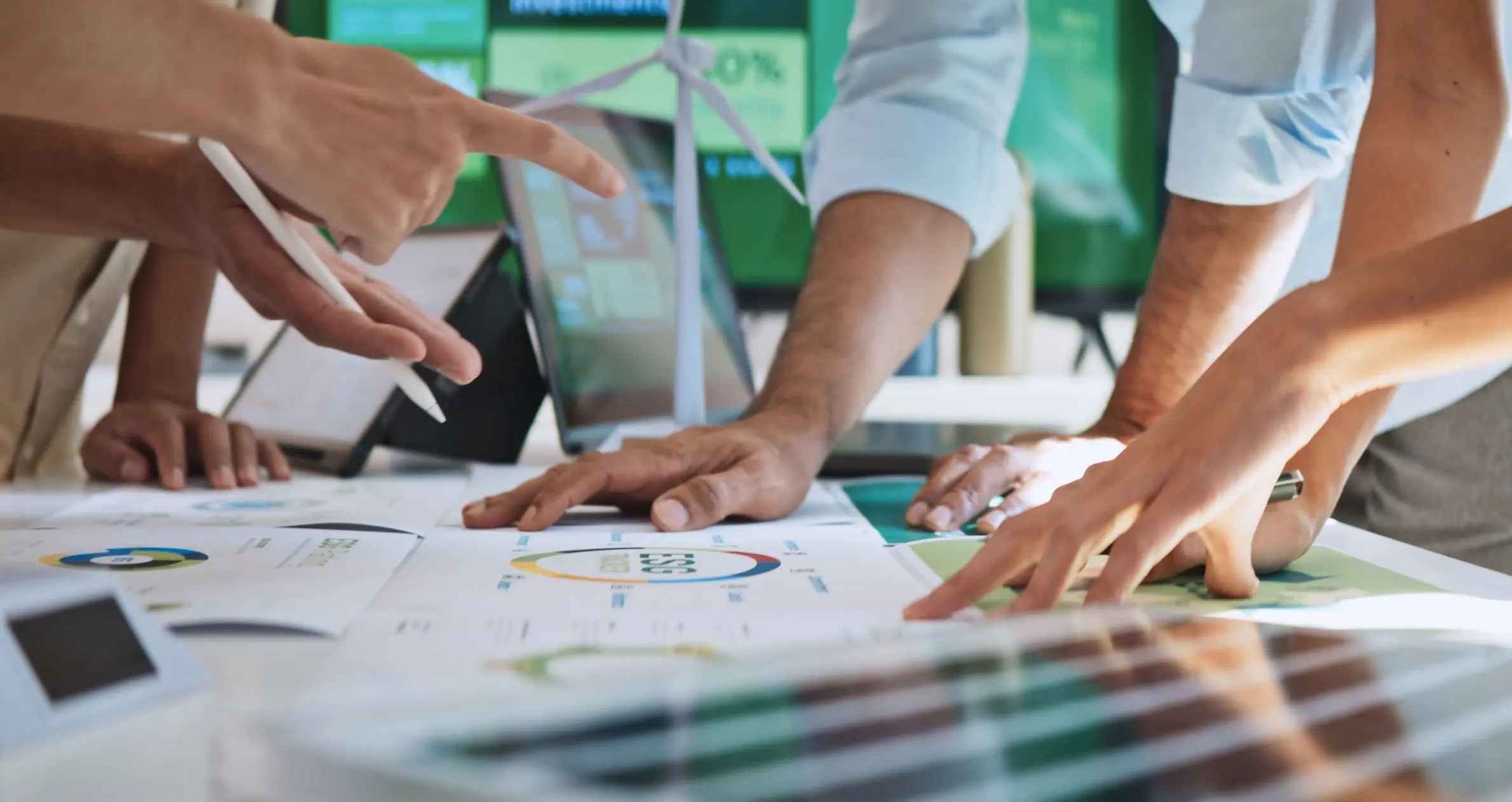 A group of people huddle around a table covered in architectural plans and charts, discussing energy-efficient designs with a wind turbine model visible in the background.