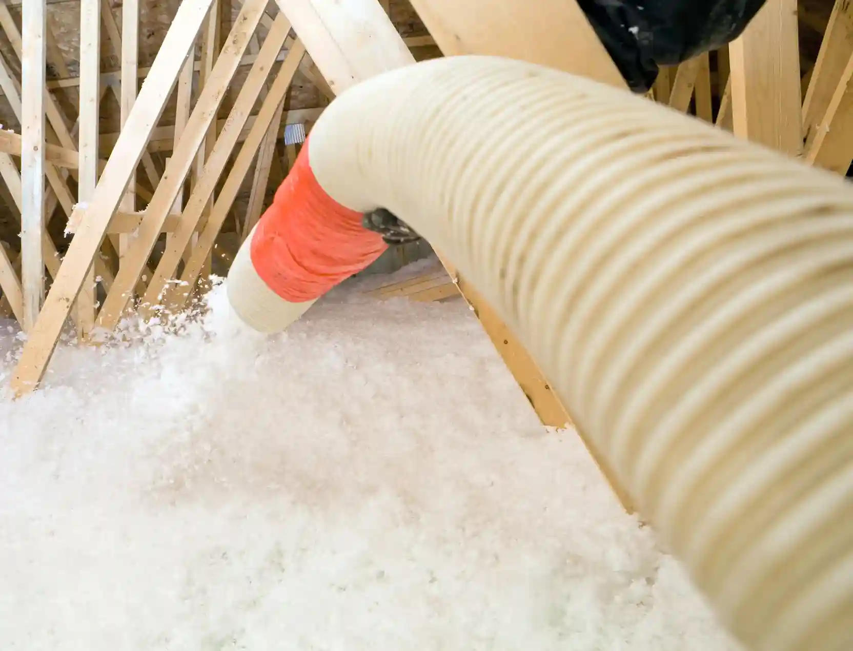 A large, thick hose with an orange band blows a cloud of white, fluffy insulation into an attic floor with wooden beams.
