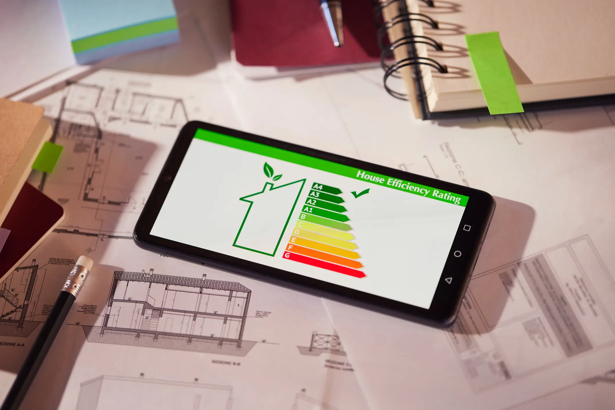 A tablet on a desk with architectural plans displays a digital house efficiency rating on a green-to-red scale, indicating a high 'A4' rating.