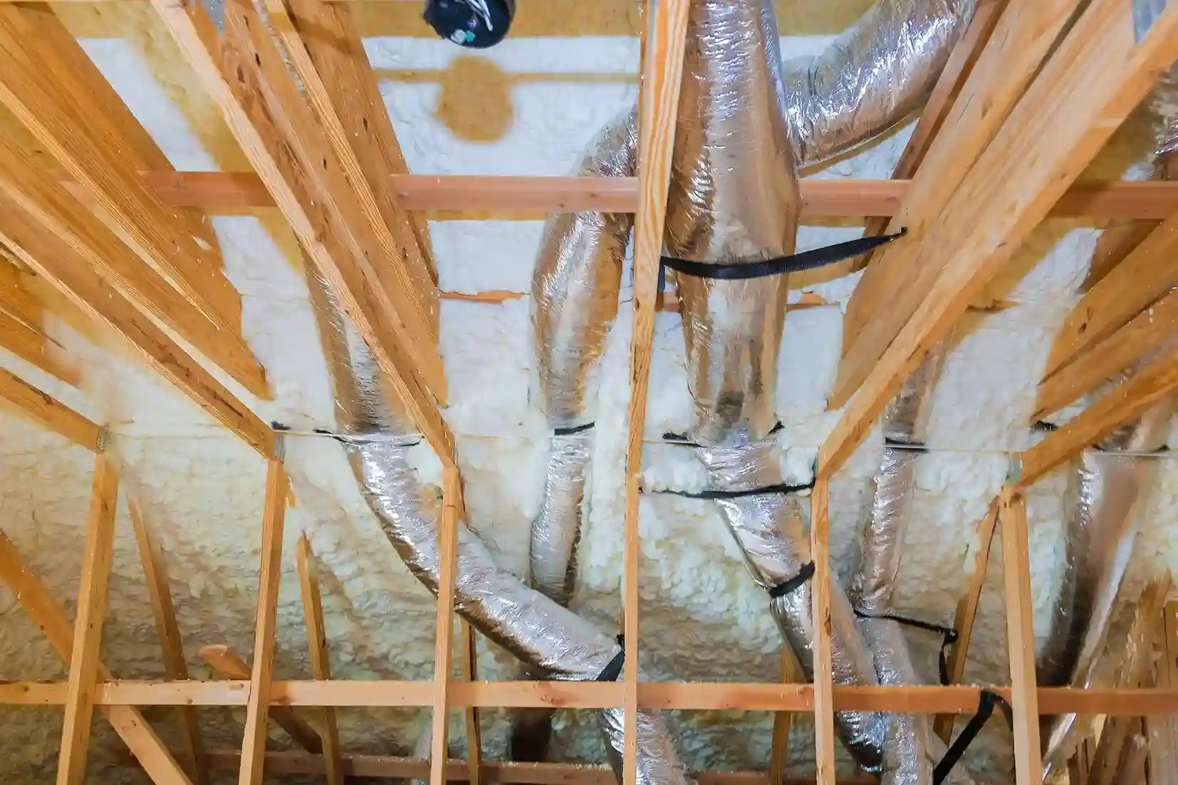 An upward view of an attic with large, silver HVAC ducts wrapped in black straps, with white blown-in insulation filling the spaces between wooden rafters.