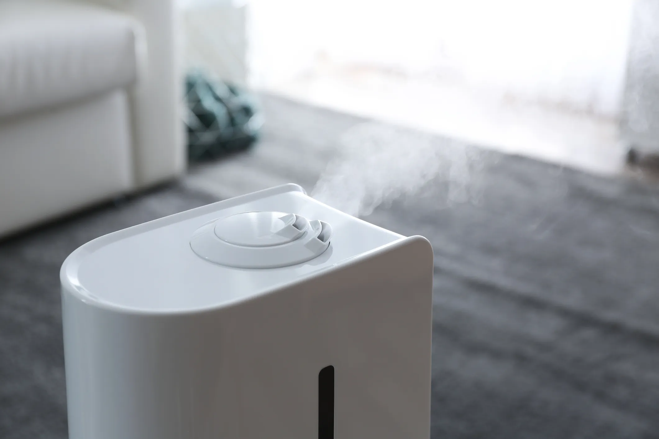 A close-up of the top of a white humidifier shows mist rising from its nozzle in a living room with a gray rug.