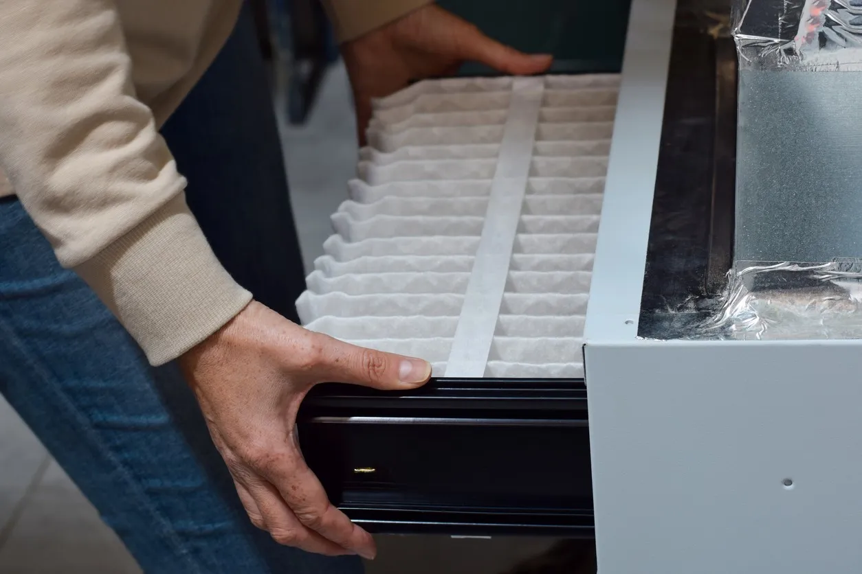 Person inserts rectangular air filter with purple edge into black furnace unit; water heater visible in background.