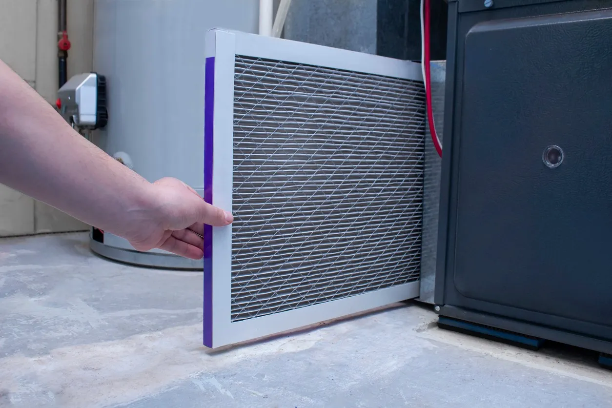 Person inserts rectangular air filter with purple edge into black furnace unit; water heater visible in background.
