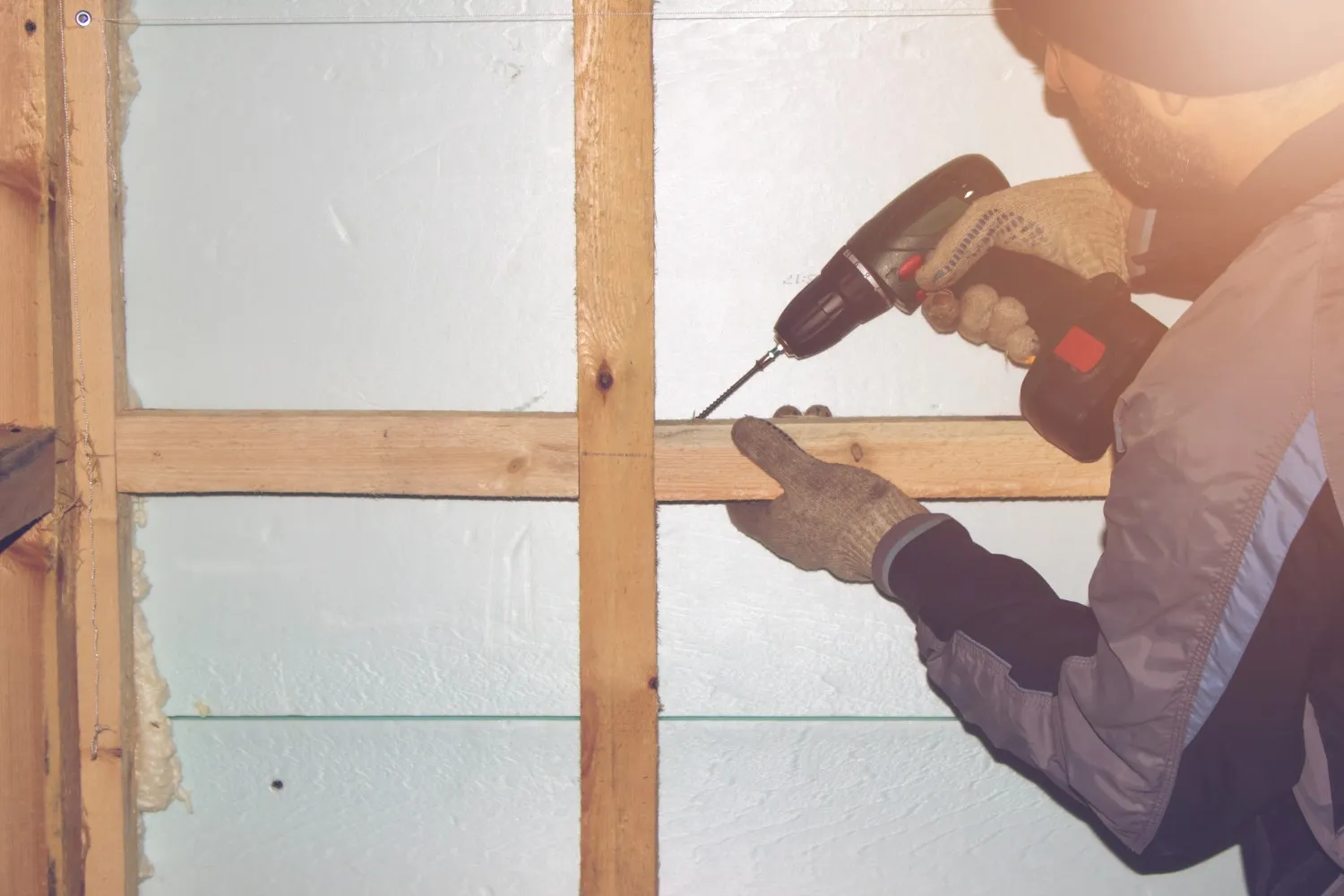 A worker using a cordless drill to secure wooden framing against an insulated wall.
