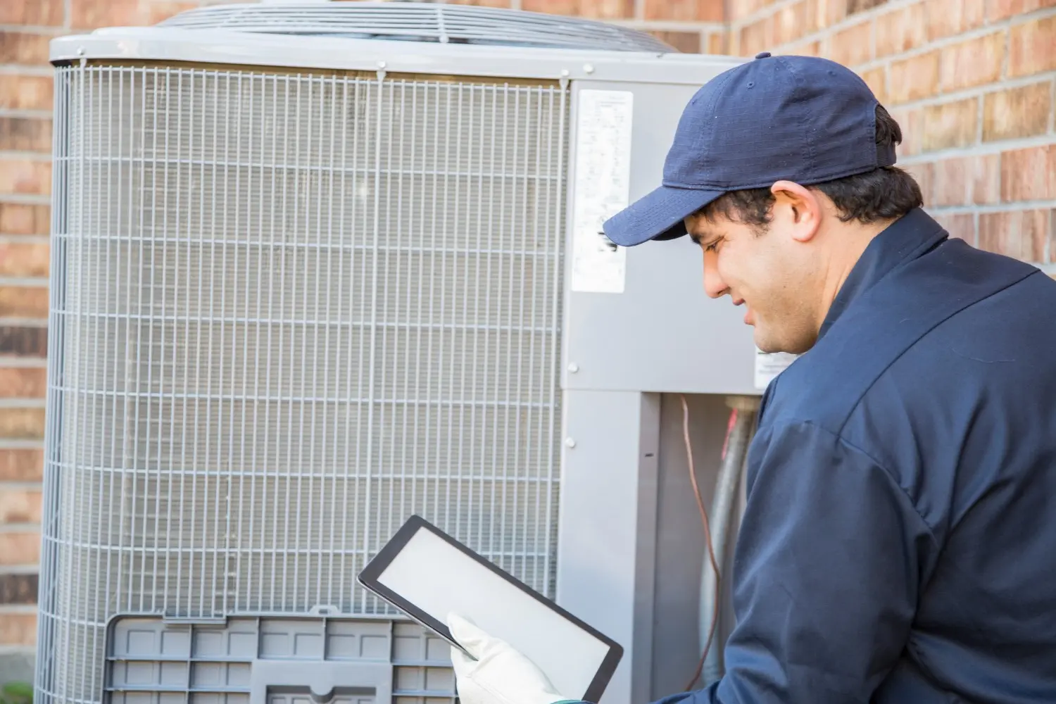 A technician in a blue cap and work shirt using a digital tablet while inspecting a residential AC unit.