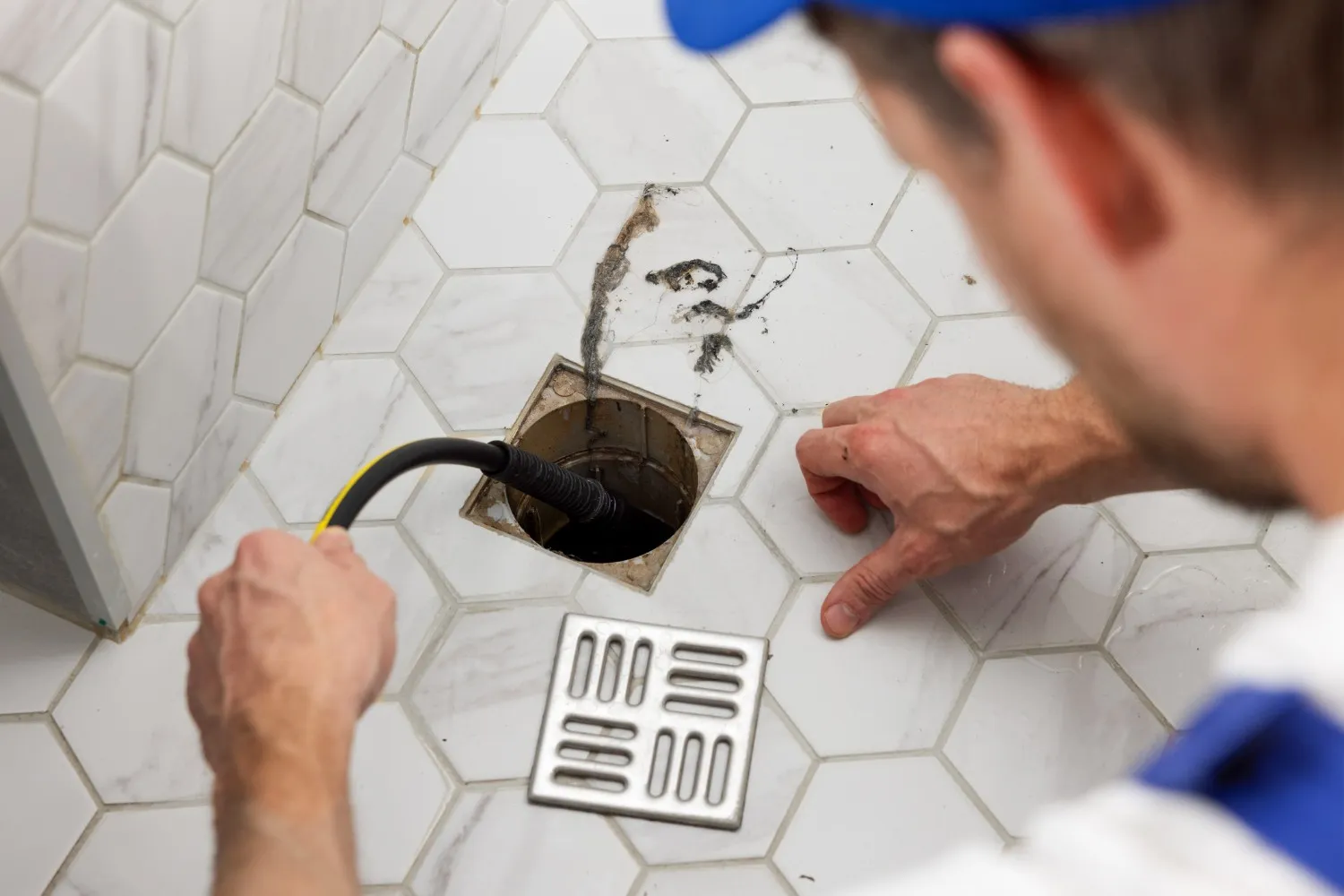 A plumber directs a black hose with high-pressure water into a shower drain opening on a white hexagonal tile floor to clear a blockage.