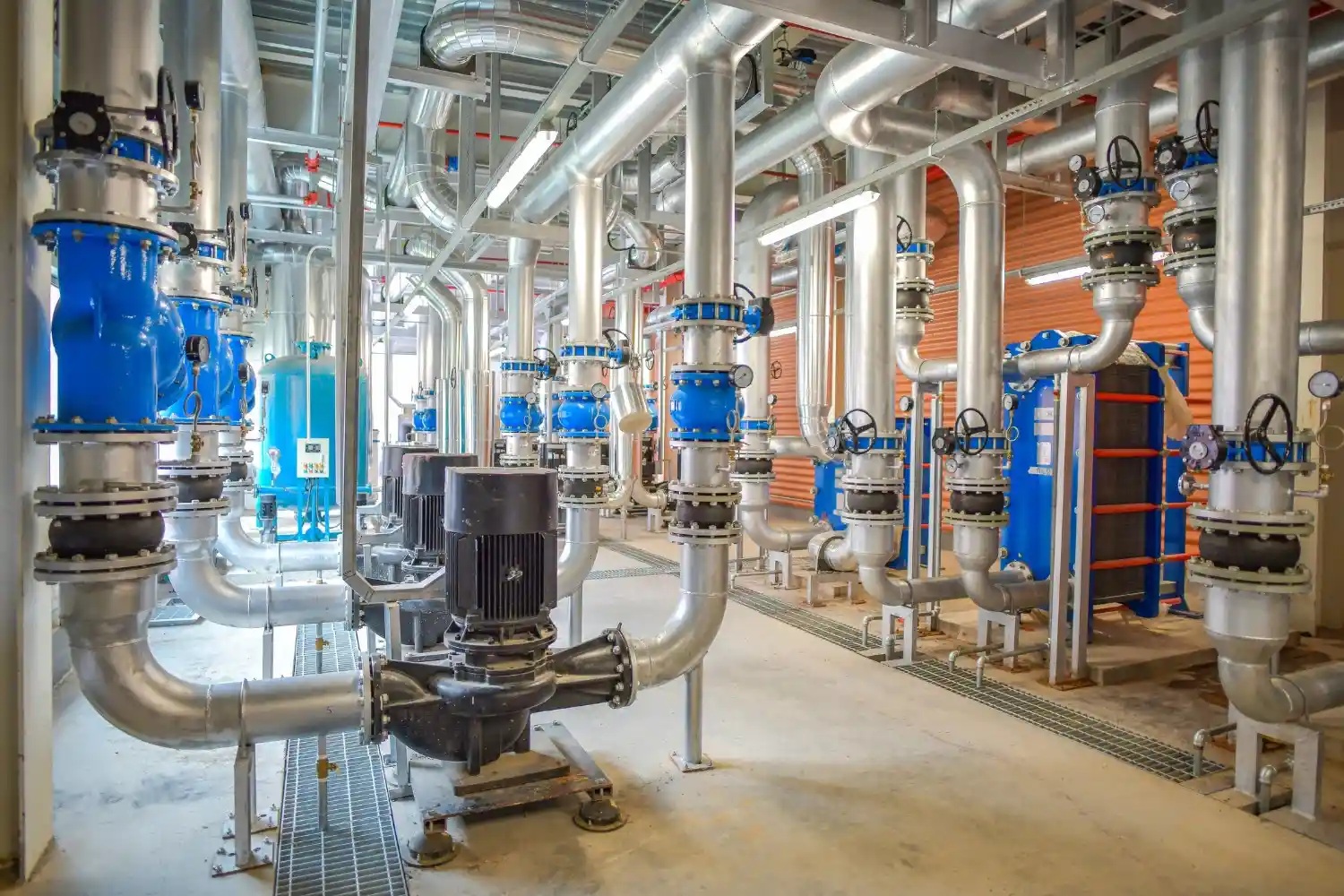 An industrial boiler room featuring a complex network of insulated silver pipes, blue valves, and large pumps.