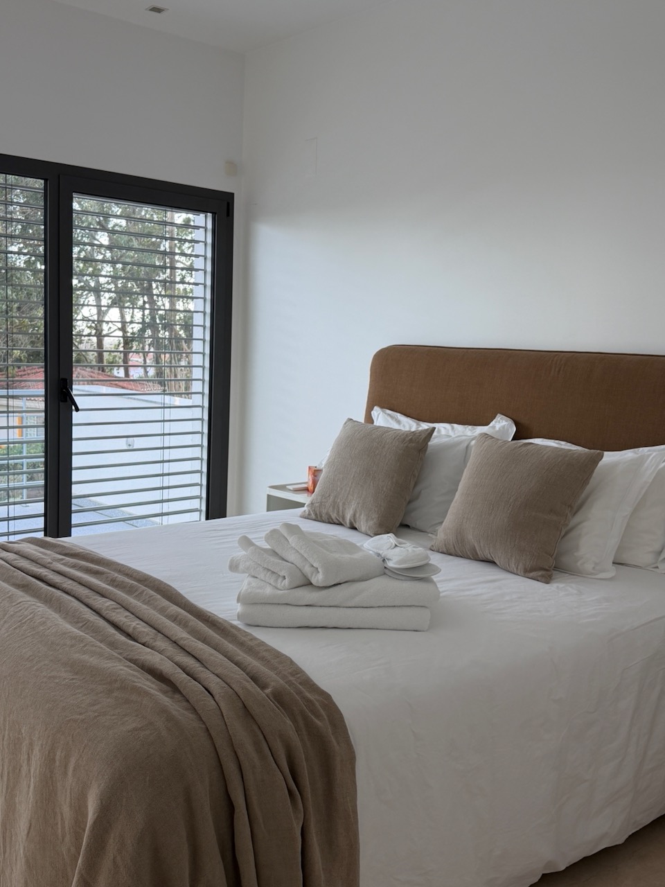A modern bedroom with a large bed featuring a brown headboard, white bed linens, two brown pillows, folded white towels, and beige slippers on the bed, next to a beige throw blanket at the foot of the bed, with a window showing outdoor trees and a building.