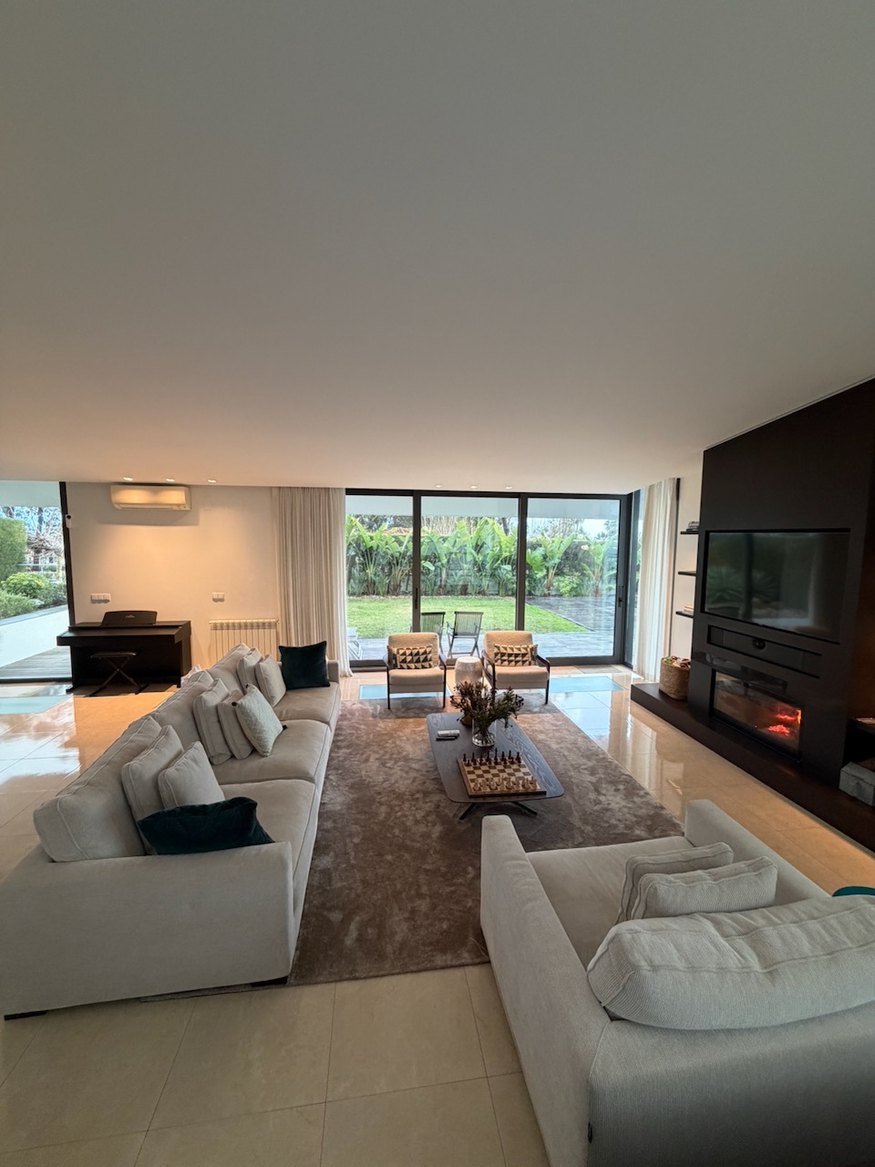 Modern living room with two large beige sofas facing a dark wood coffee table with a chess set and vase, two armchairs near large glass sliding doors overlooking a garden, a black TV mounted above a fireplace on the right, and a piano on the left.