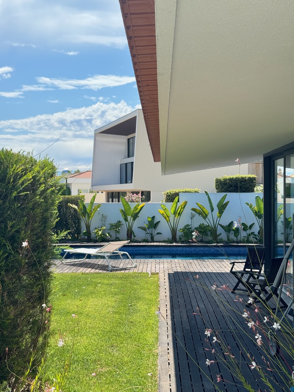 Modern backyard with green lawn, pool, and wooden deck featuring black chairs under an overhanging roof, surrounded by white walls and plants. A white sun lounger sits near the pool. A contemporary house with large windows and a pitched roof is visible beyond the backyard fence.