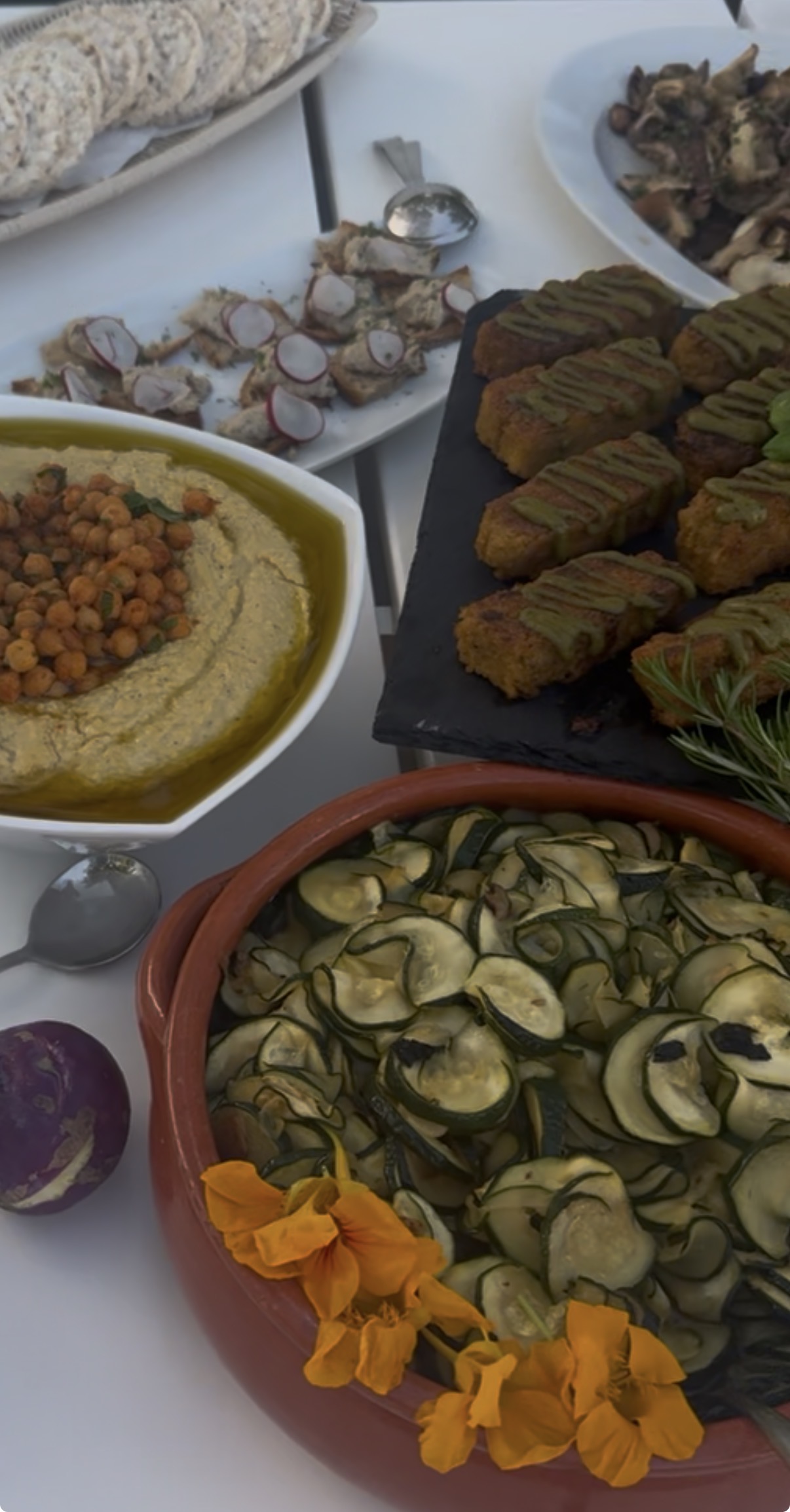 A spread of assorted food dishes on a white table including sliced bread, hummus topped with chickpeas and olive oil, croquettes drizzled with green sauce on a slate platter, roasted zucchini slices in a terracotta bowl garnished with orange edible flowers, and small bread slices topped with radish slices and herbs.