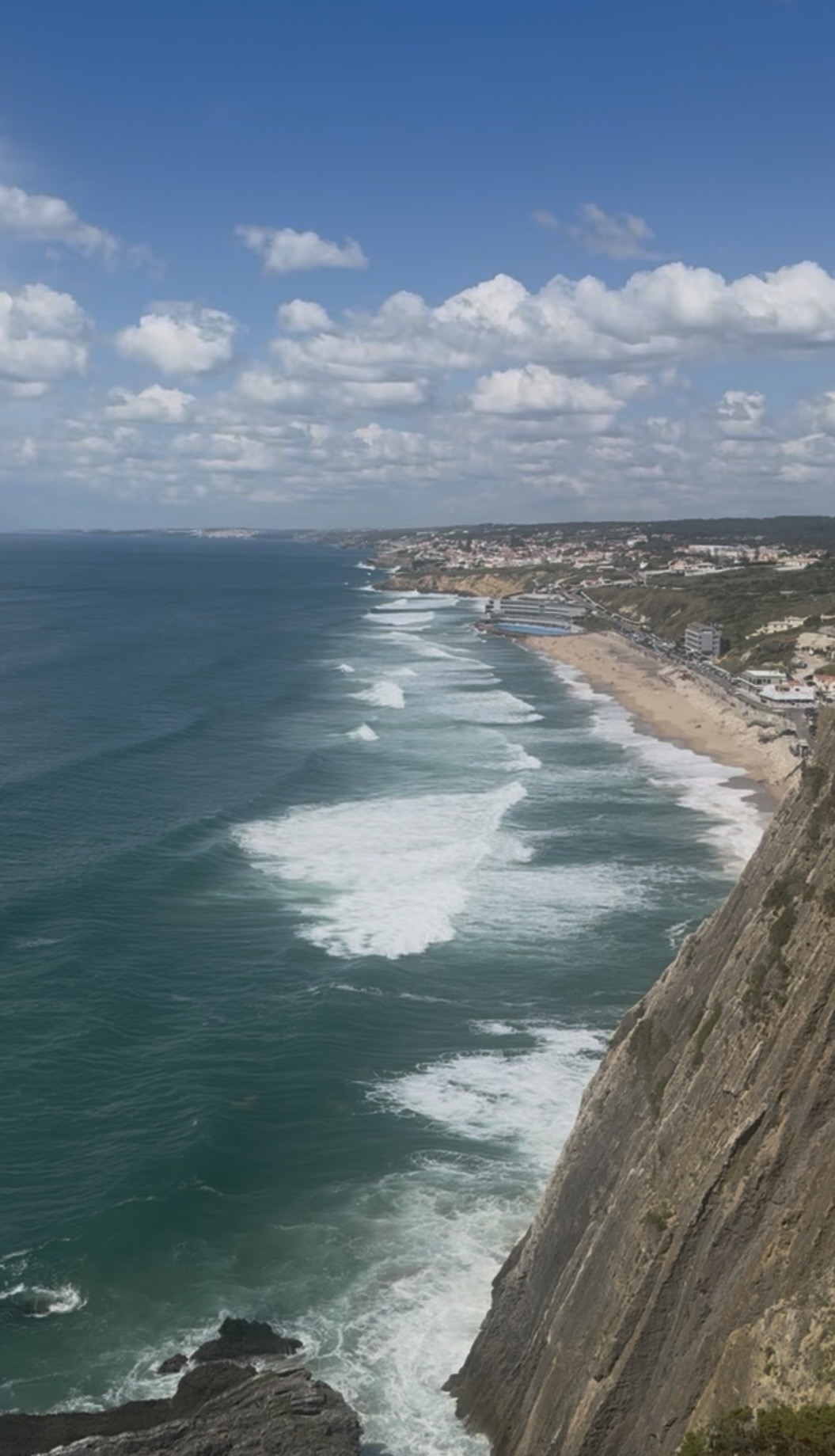 View from a steep cliff overlooking a coastal town with a sandy beach and waves crashing along the shoreline under a partly cloudy blue sky.