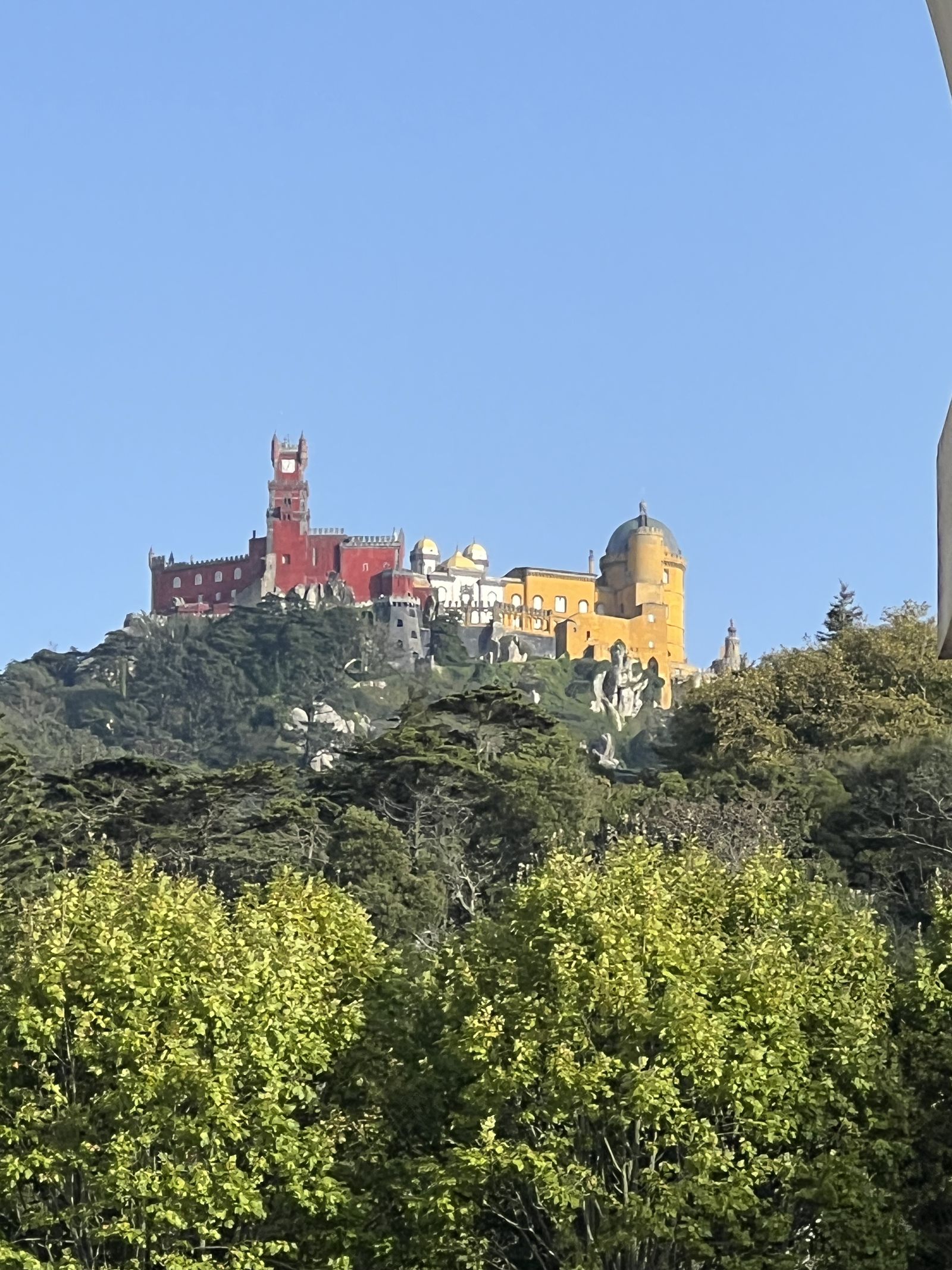 Colorful Pena Palace atop a lush green hill under a clear blue sky, with red, yellow, and white sections visible.