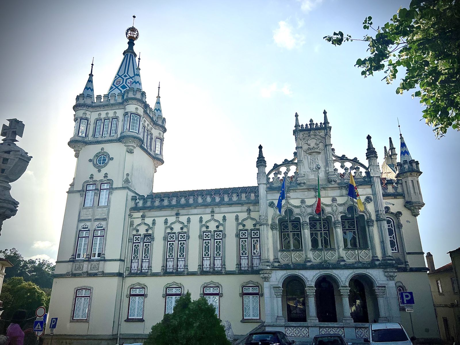Historic ornate building with two tall towers topped by blue-and-white patterned spires and intricate stonework, featuring multiple windows and arched entrance, with flags displayed on the facade under a partly cloudy sky.
