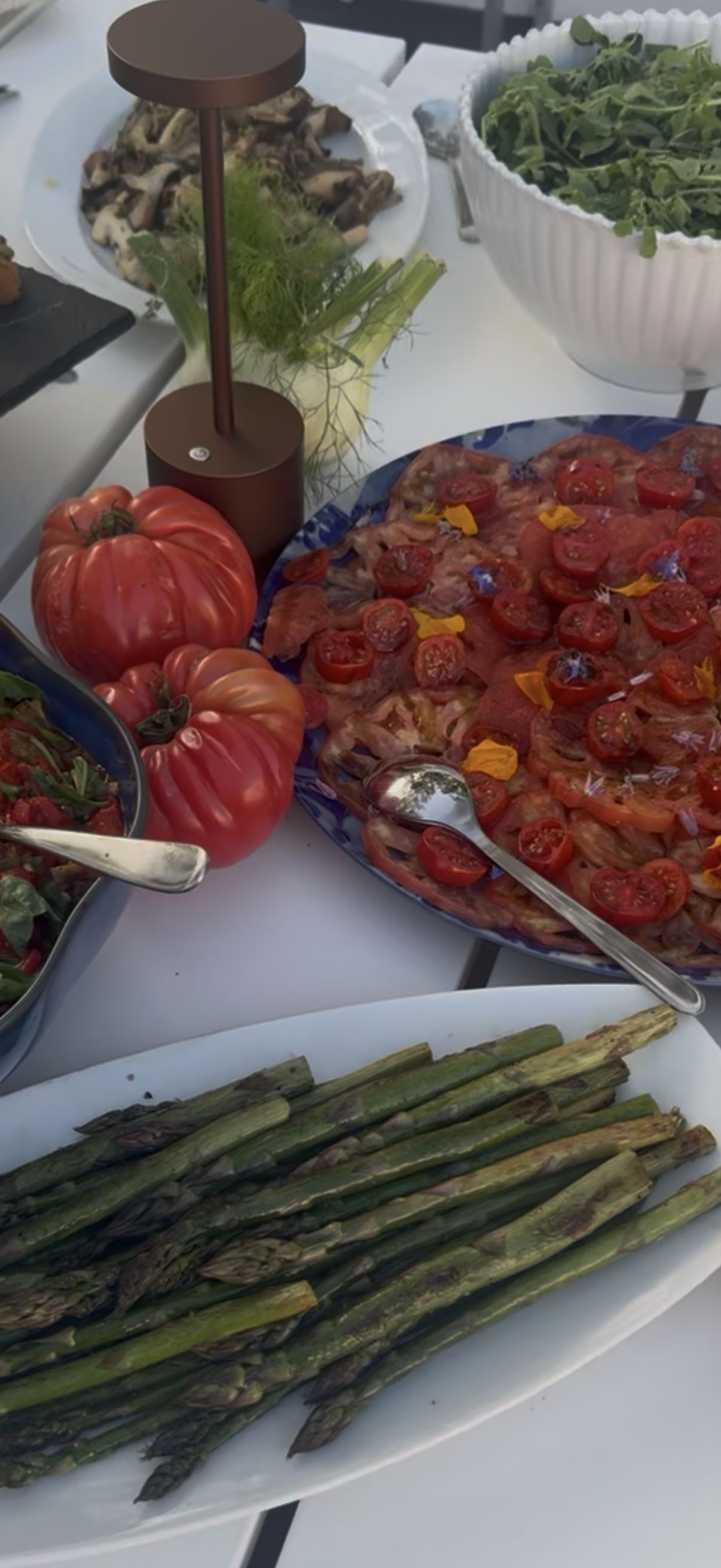 Plated food on a white table includes grilled asparagus on a white serving dish, heirloom tomatoes, a plate of sliced tomatoes topped with cherry tomato halves and edible flowers, a bowl of leafy greens, and a plate of sautéed mushrooms with fennel on the side.