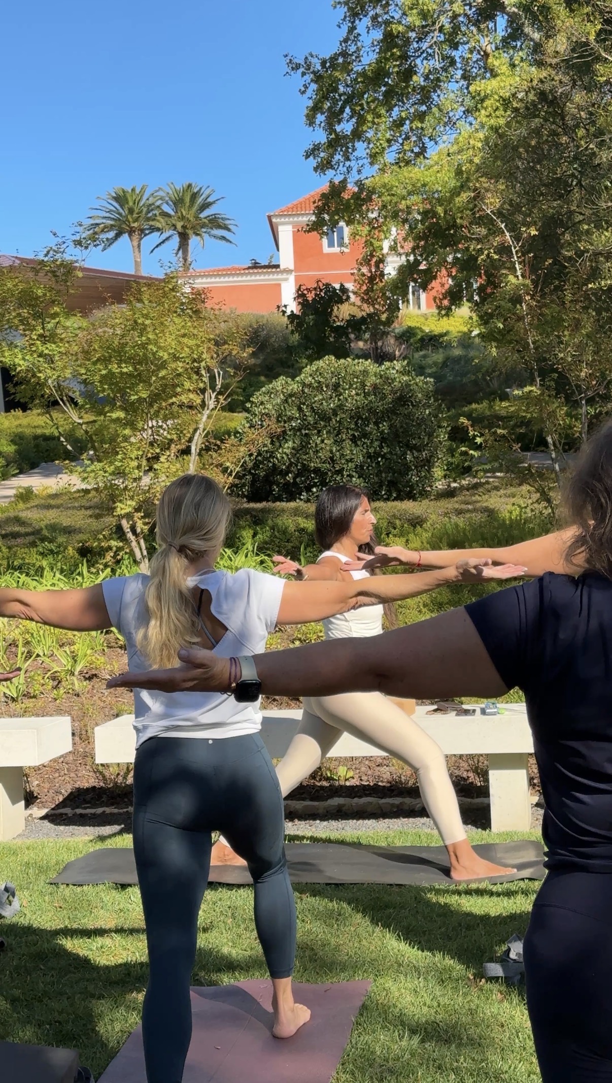 Three women practicing yoga outdoors on mats in a garden under clear blue sky, performing a standing balancing pose with arms extended.